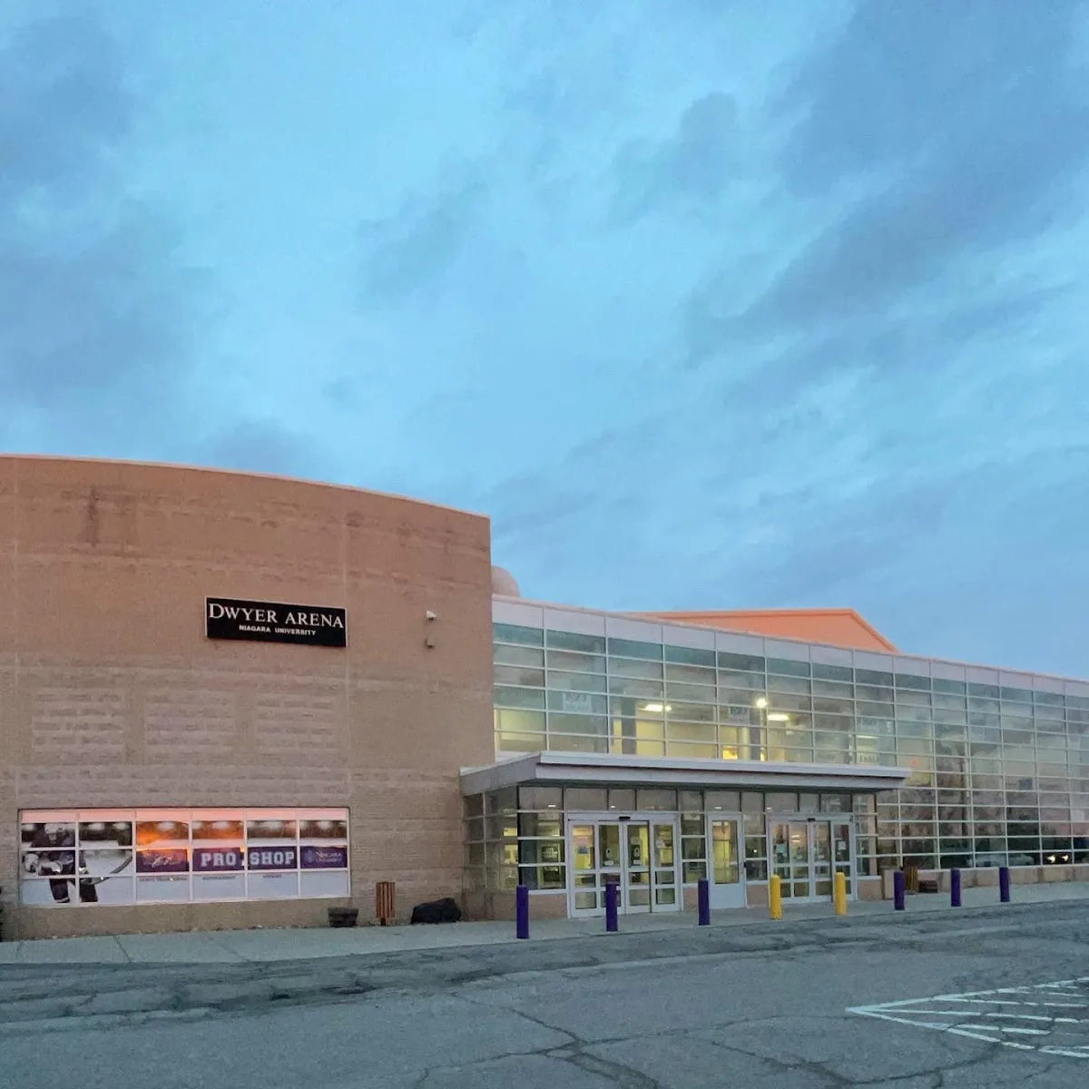 The tan brick Dwyer Arena front with its DWYER ARENA sign, a glass entrance and the pro shop windows, seen from the parking lot at dusk. — Dwyer Arena