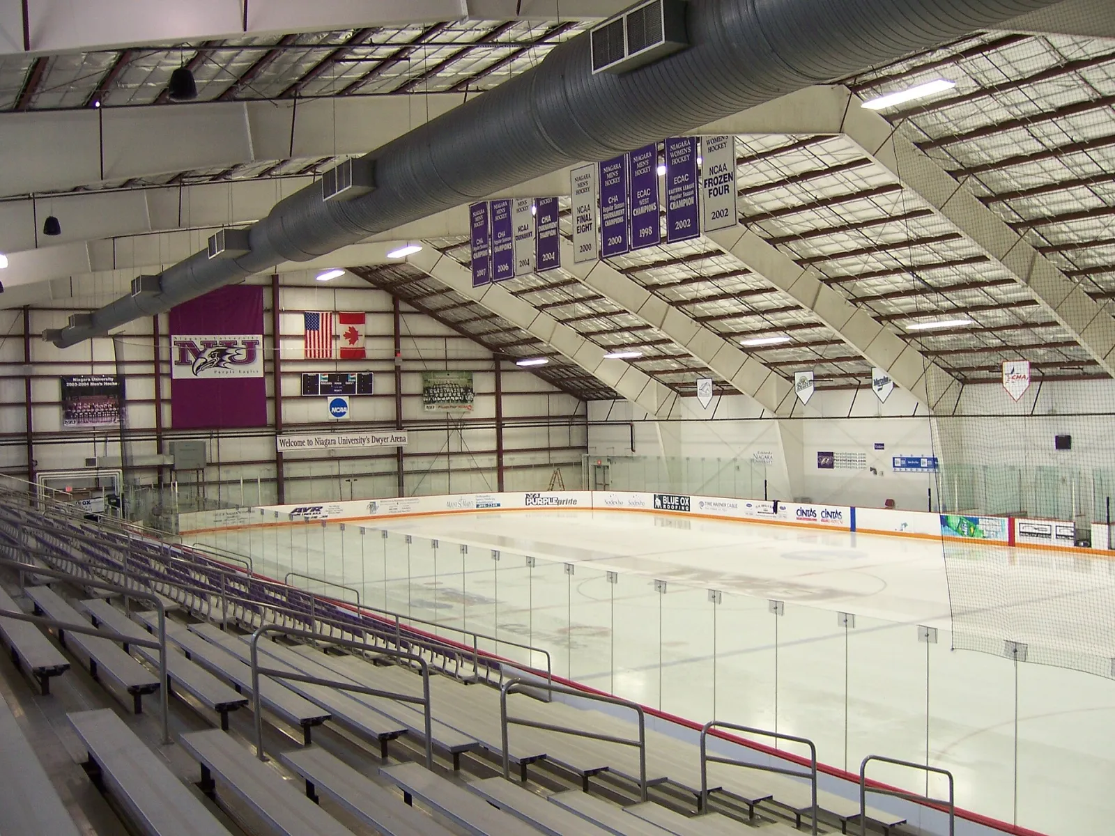 Dwyer Arena's main rink seen from the upper stands, with purple Niagara banners and flags hanging from the steel roof over empty ice. — Dwyer Arena
