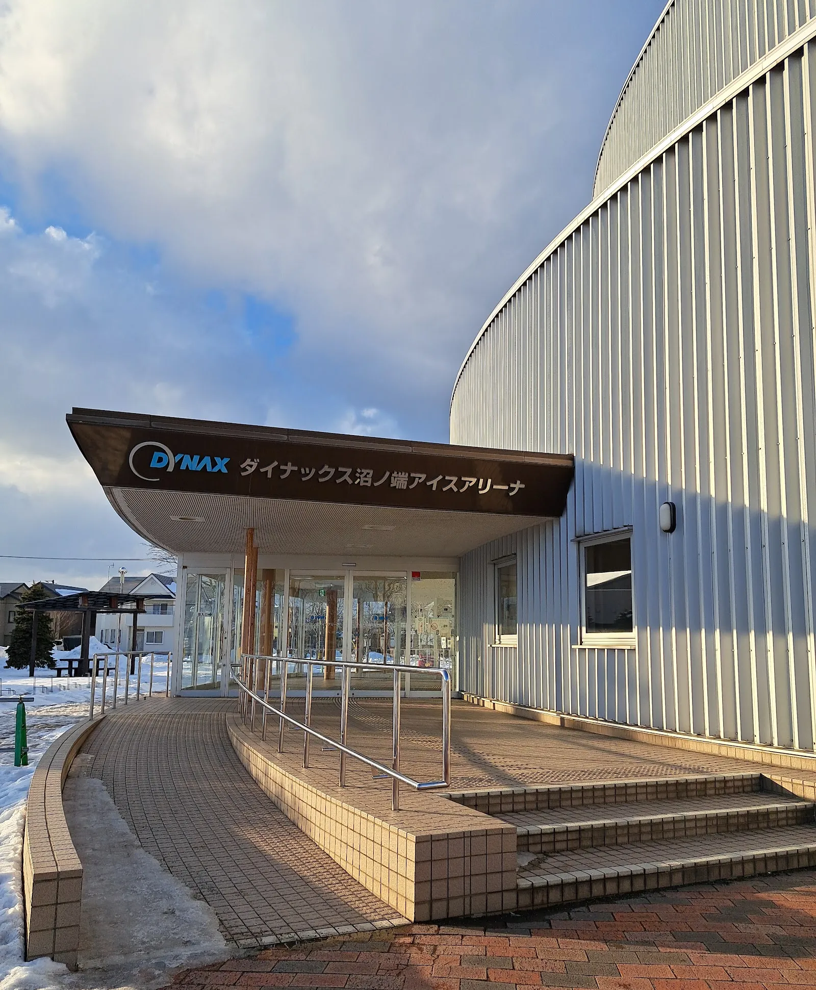 冬の苫小牧にあるDYNAXぬまのはたアイスアリーナ。曲線を描く白い金属張りの建物で、入口の庇の上に名称の看板があり、階段とレンガ舗装が見えます。 — Dynax Numanohata Ice Arena