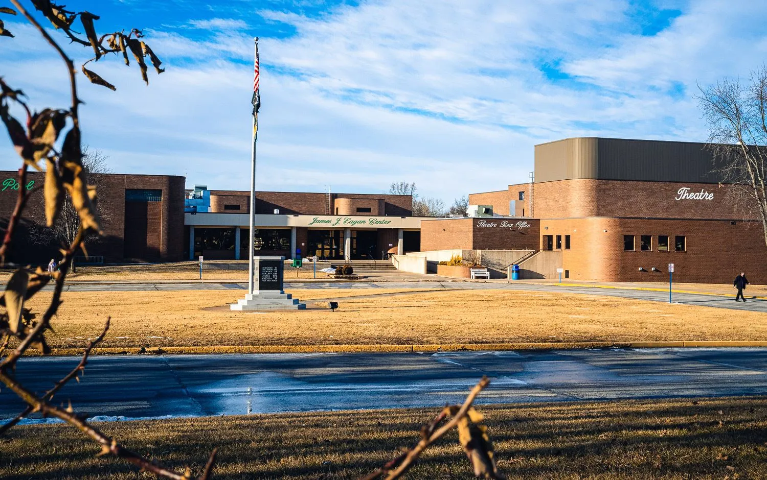 El bajo edificio de ladrillo del James J. Eagan Center del otro lado de un césped con un asta de bandera y un monumento, con el ala del teatro a la derecha. — James J. Eagan Center Ice Rink