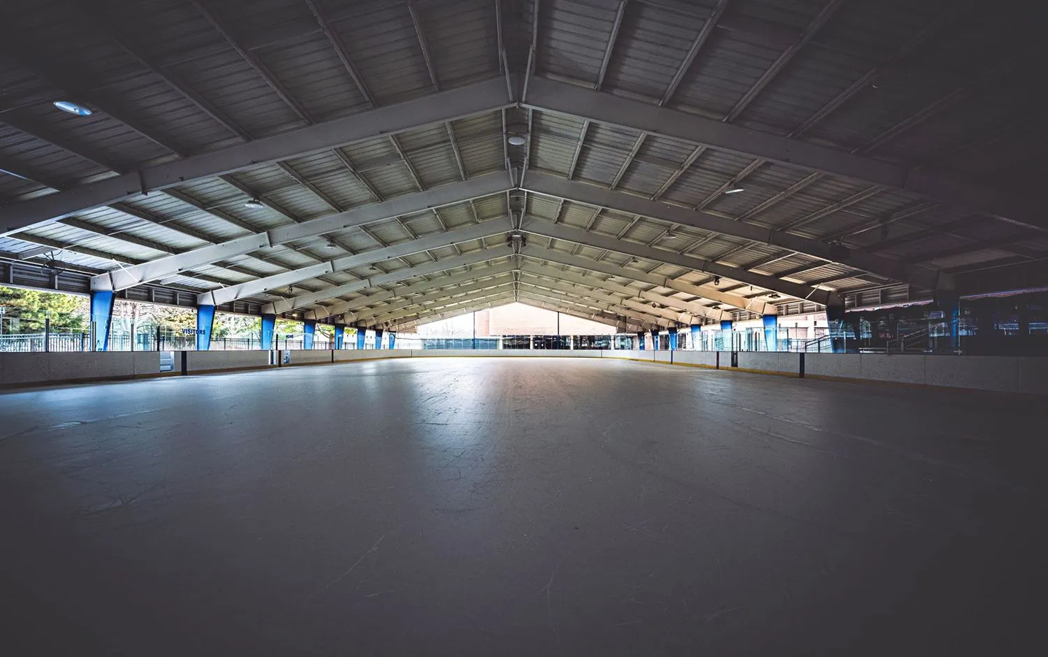 La pista de hielo vacía bajo el largo techo con estructura de acero, con los costados abiertos y las bardas a lo largo de todo el pabellón. — James J. Eagan Center Ice Rink