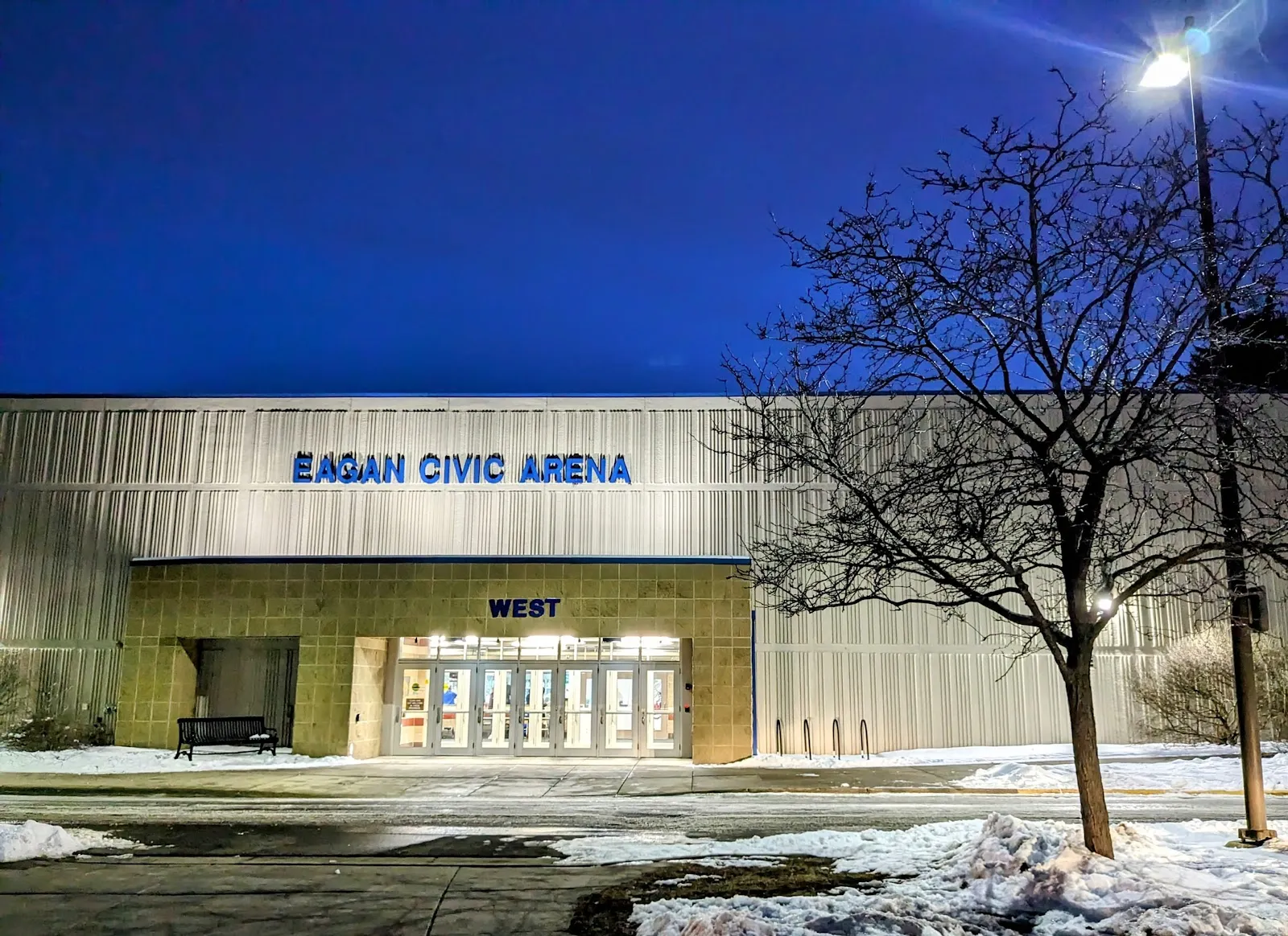 The West entrance at dusk: the EAGAN CIVIC ARENA sign across the metal facade above a stone entry with glass doors, snow on the lawn in front. — Eagan Civic Arena