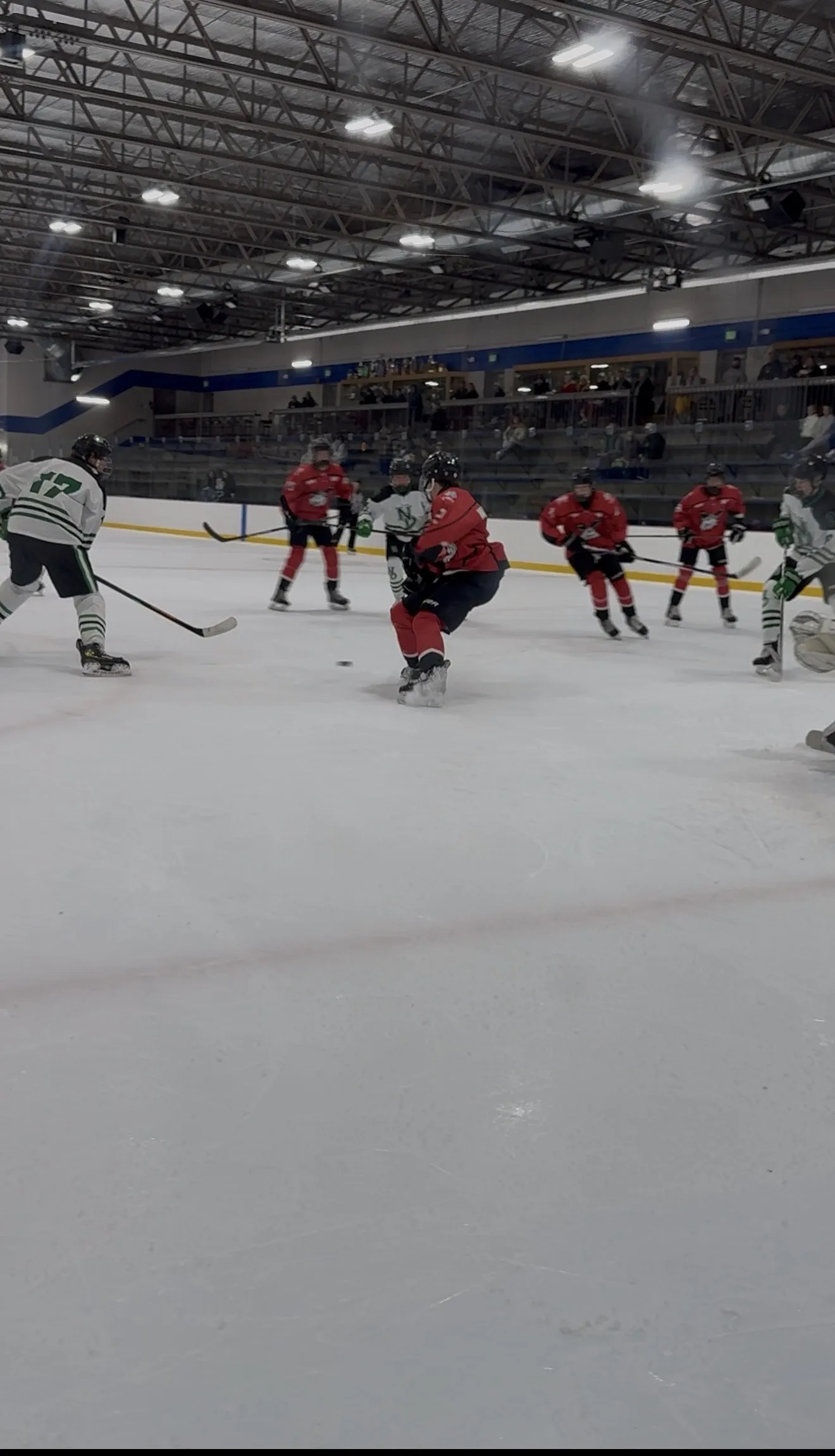 A game seen from behind the glass, players spread across a dark-floored sheet under a lit steel ceiling with a goal at the right. — Eagan Civic Arena
