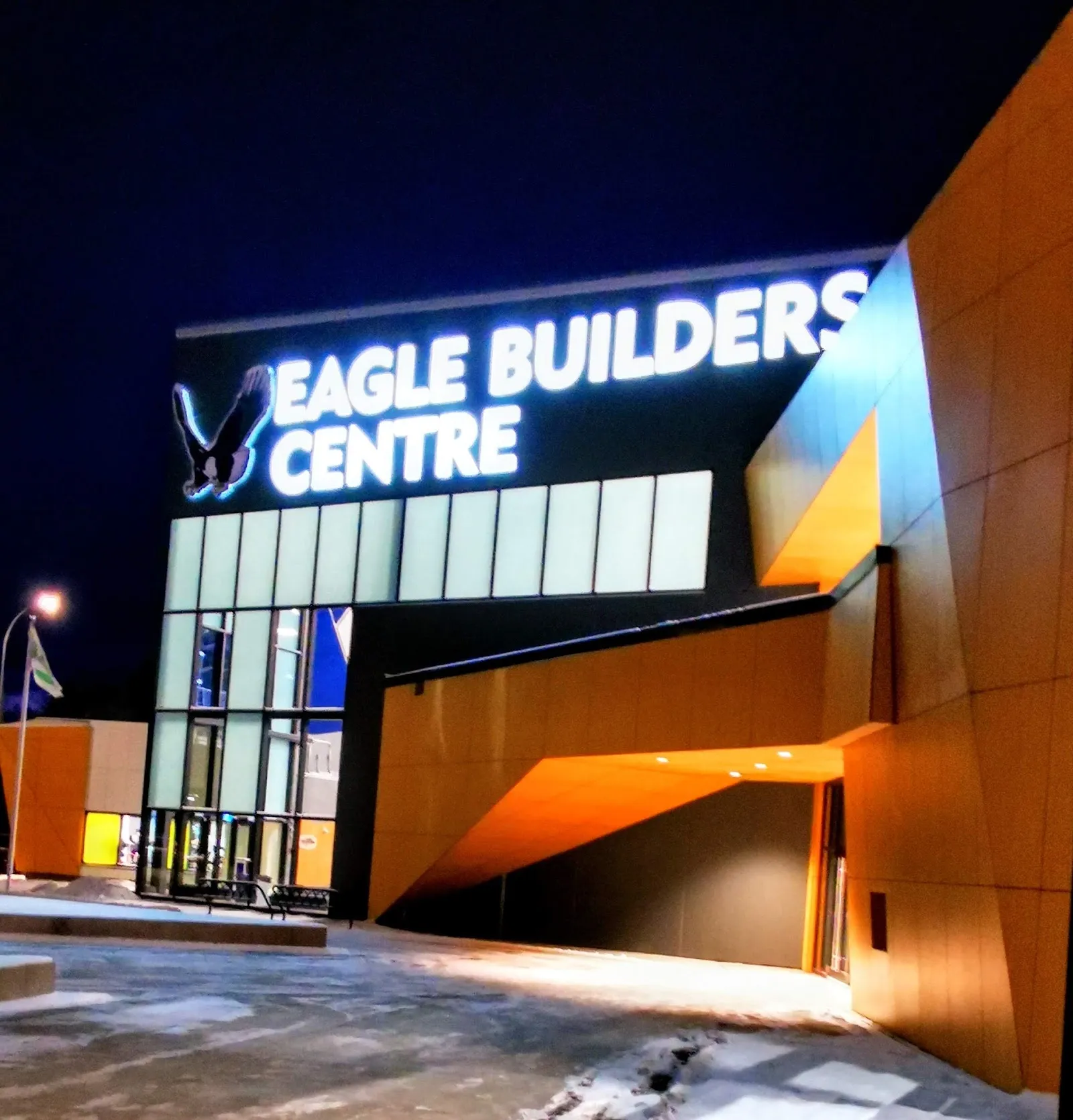 The glazed entrance and orange-clad front of the Eagle Builders Centre at night, with the illuminated Eagle Builders Centre sign and eagle logo. — Eagle Builders Centre