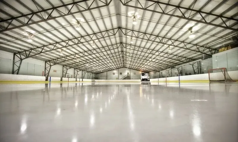 Una larga pista vacía bajo un techo de armaduras de acero con una máquina alisadora estacionada en el extremo opuesto y las luces reflejadas en el hielo. — Eagles Ice Arena