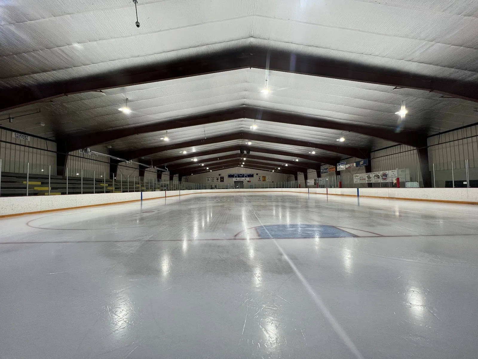An empty sheet seen from the end of the boards, white ice with a blue centre line under a red-trussed roof with a light grey ceiling. — East Alton Ice Arena