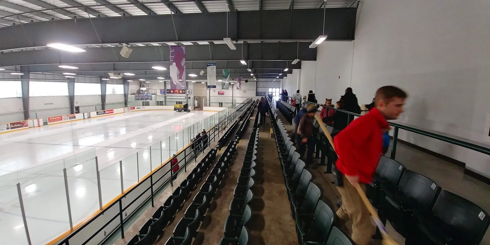 Una vista amplia de la pista desde las gradas altas, con jugadores repartidos por el hielo y hileras de asientos morados a lo largo del lado cercano. — East Bayfield Community Centre