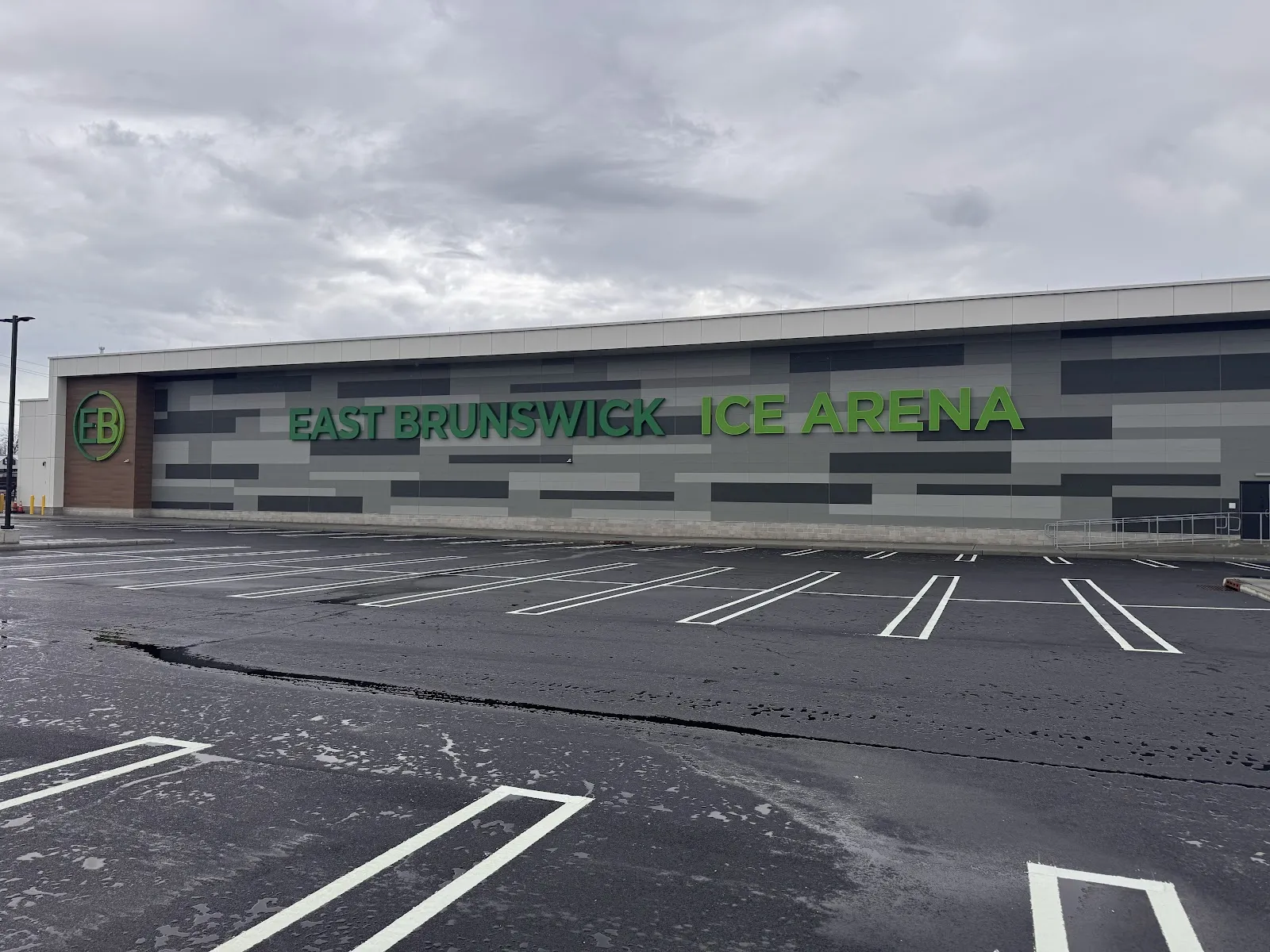 El largo muro gris y verde de East Brunswick Ice Arena con su nombre y el logotipo EB, visto al otro lado del estacionamiento recién marcado — East Brunswick Ice Arena