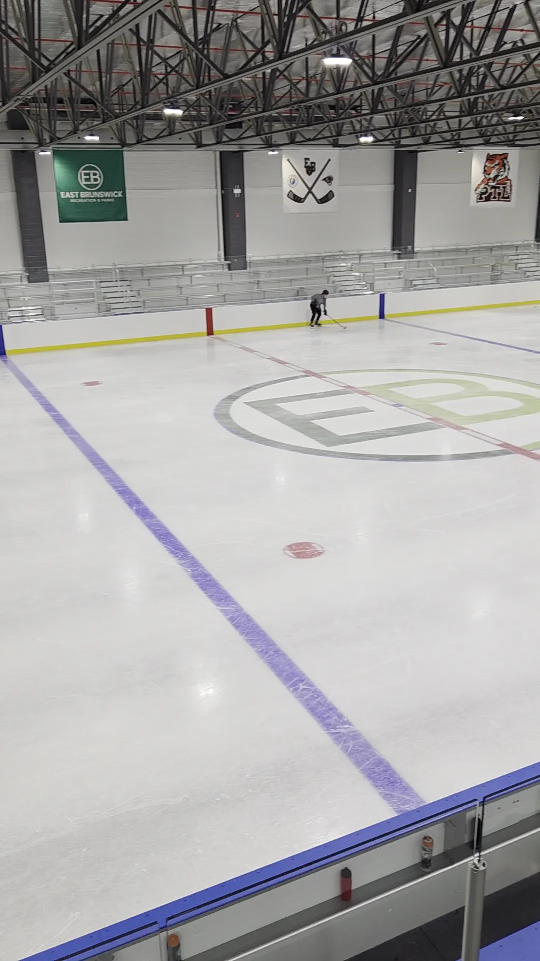 La nueva pista de East Brunswick Ice Arena con el logotipo EB en el centro del hielo y un patinador a lo lejos bajo las armaduras negras de acero del techo — East Brunswick Ice Arena
