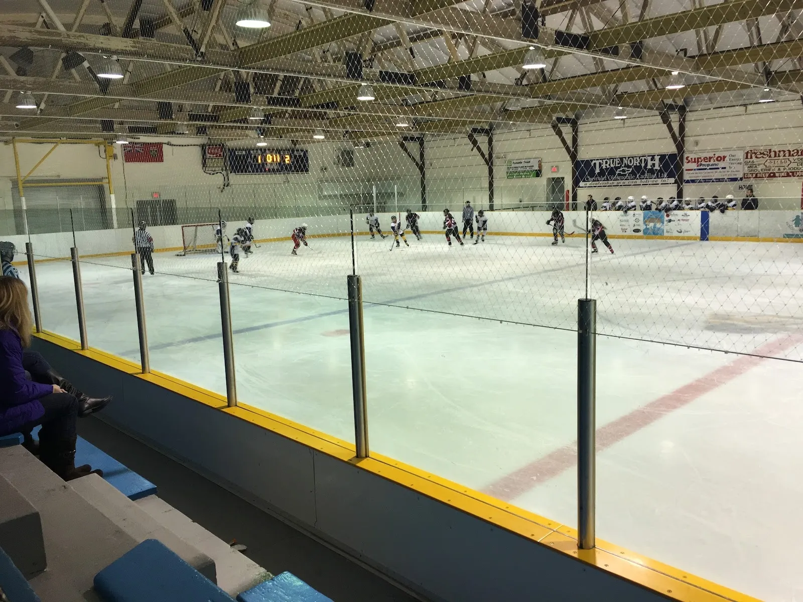 A wide view along a clear glass rail of a long sheet, a few players spread across the ice and a yellow boards line below. — East Ferris Community Centre (Astorville)
