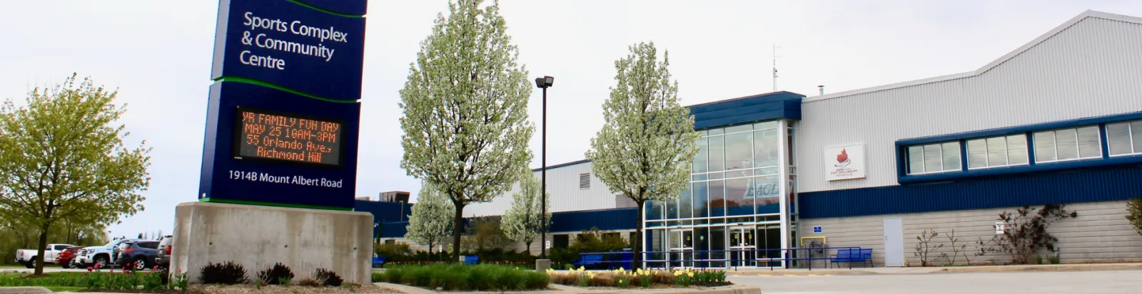 El complejo bajo, azul y blanco, detrás de su letrero junto a la carretera, que dice Sports Complex & Community Centre, 1914B Mount Albert Road. — East Gwillimbury Sports Complex