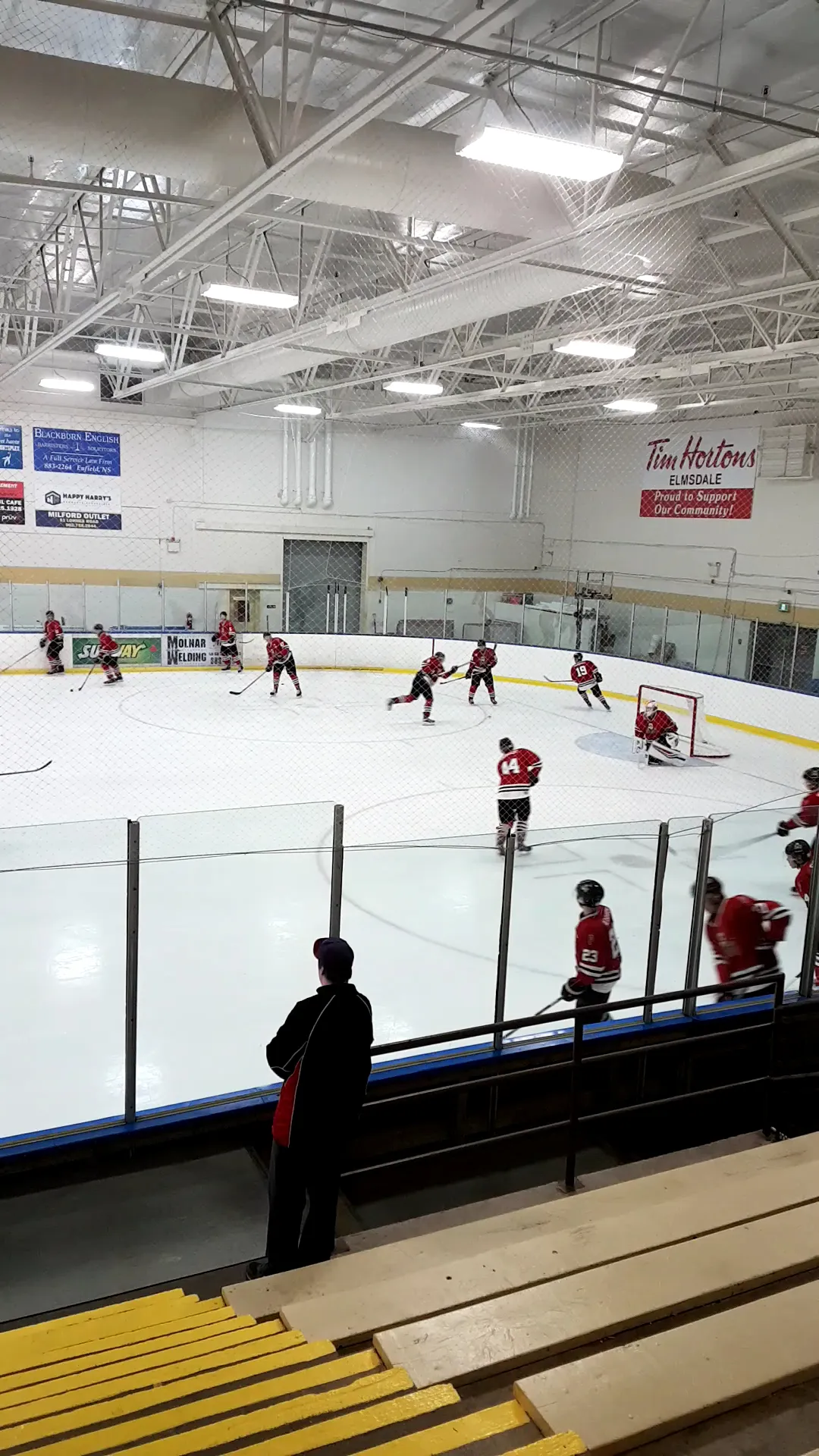 A hockey game on the ice at the East Hants Sportsplex, with sponsor boards and a Tim Hortons banner on the far wall and the steel roof trusses above. — East Hants Sportsplex