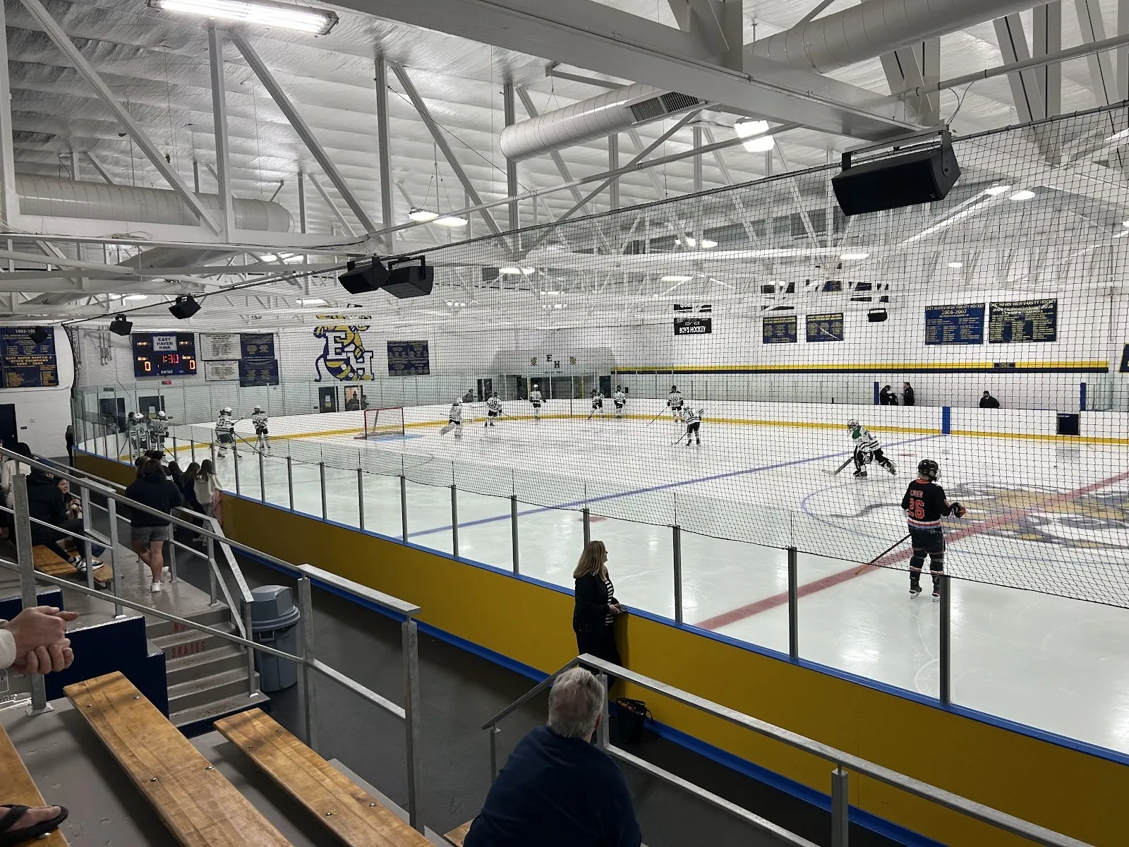 Un partido visto desde las gradas, con jugadores repartidos por una pista amplia bajo un techo de armaduras blancas y un barandal amarillo a lo largo de las vallas cercanas. — East Haven Ice Rink