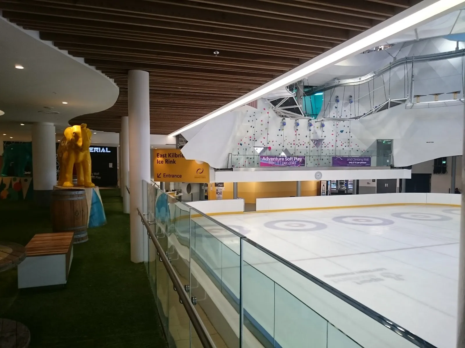 The rink's public frontage inside the Olympia Shopping Centre: the ice seen from the mall balcony behind a glass screen, with the yellow 'East Kilbride Ice Rink - Entrance' sign. The rink has no street-facing front of its own. — East Kilbride Ice Rink
