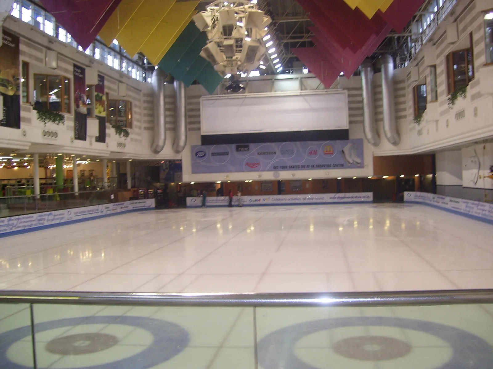 The ice surface with curling targets, seen from the viewing level inside the Olympia centre under its coloured ceiling banners. — East Kilbride Ice Rink