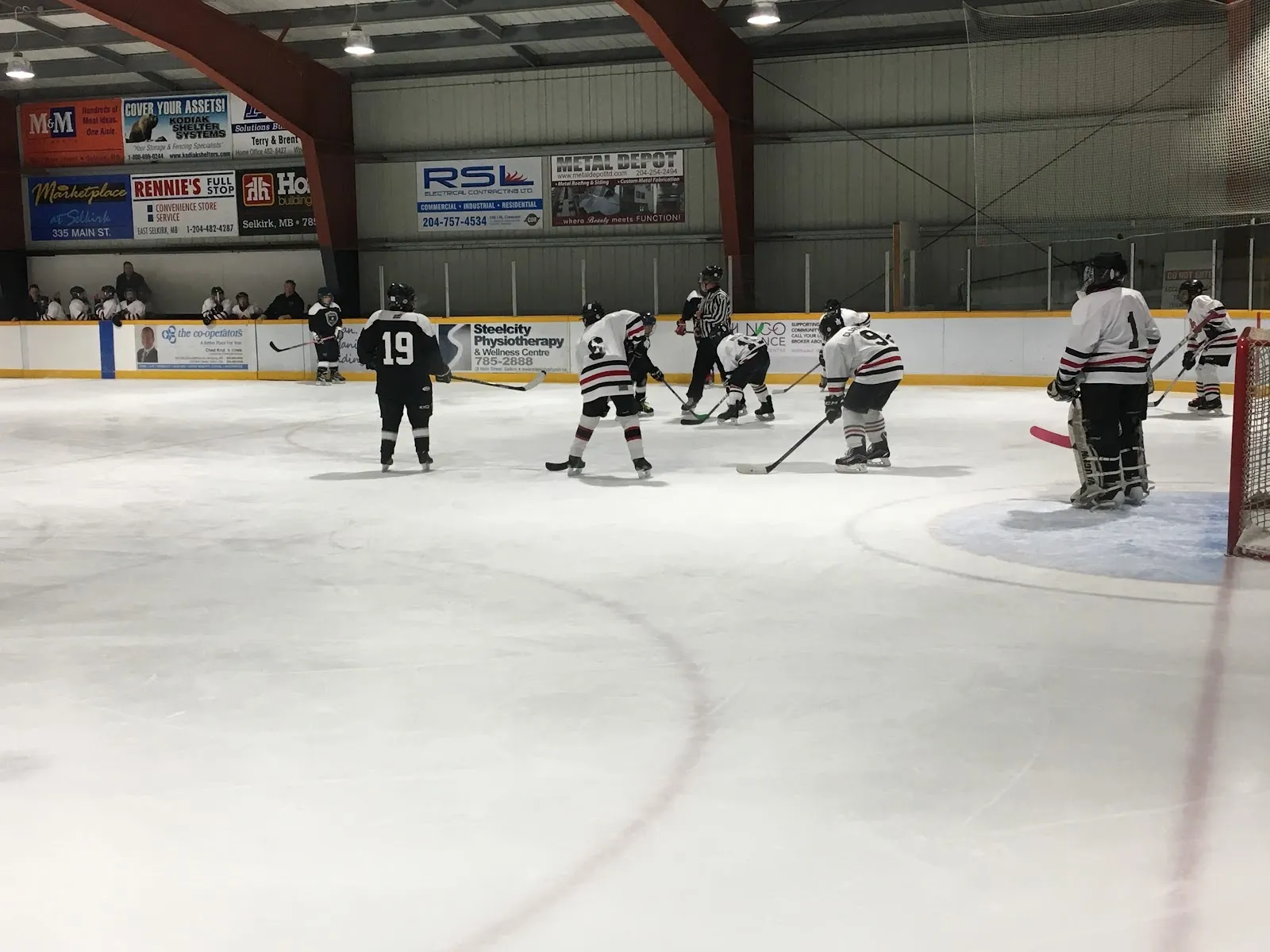 A faceoff during a game on the ice at East Selkirk Arena, with sponsor boards and red steel arches along the far wall. — East Selkirk Arena