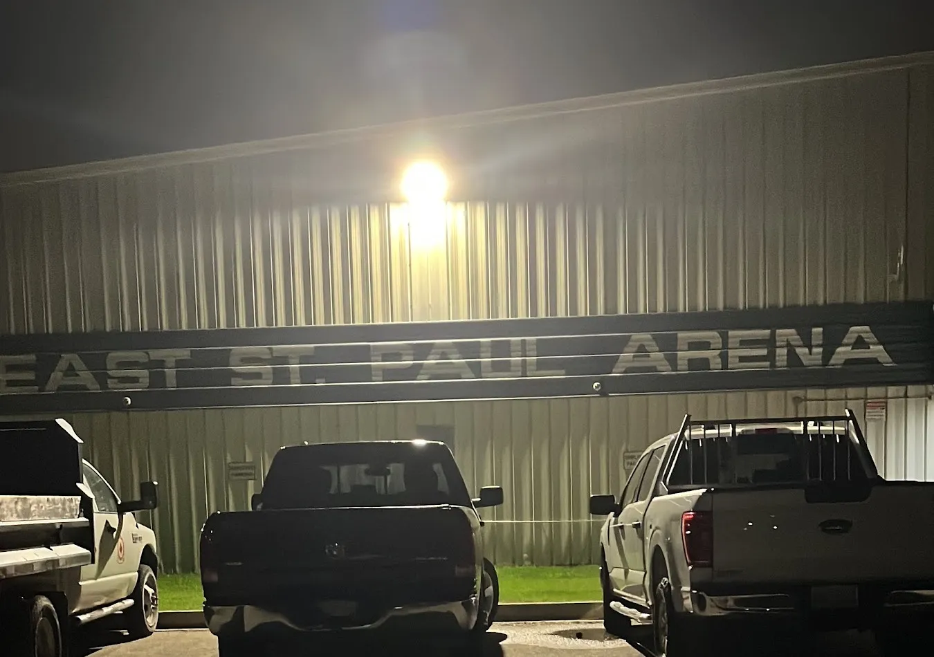 La East St. Paul Arena revestida de metal, de noche, con su nombre en la pared bajo una luz brillante y camionetas pickup estacionadas al frente. — East St. Paul Arena