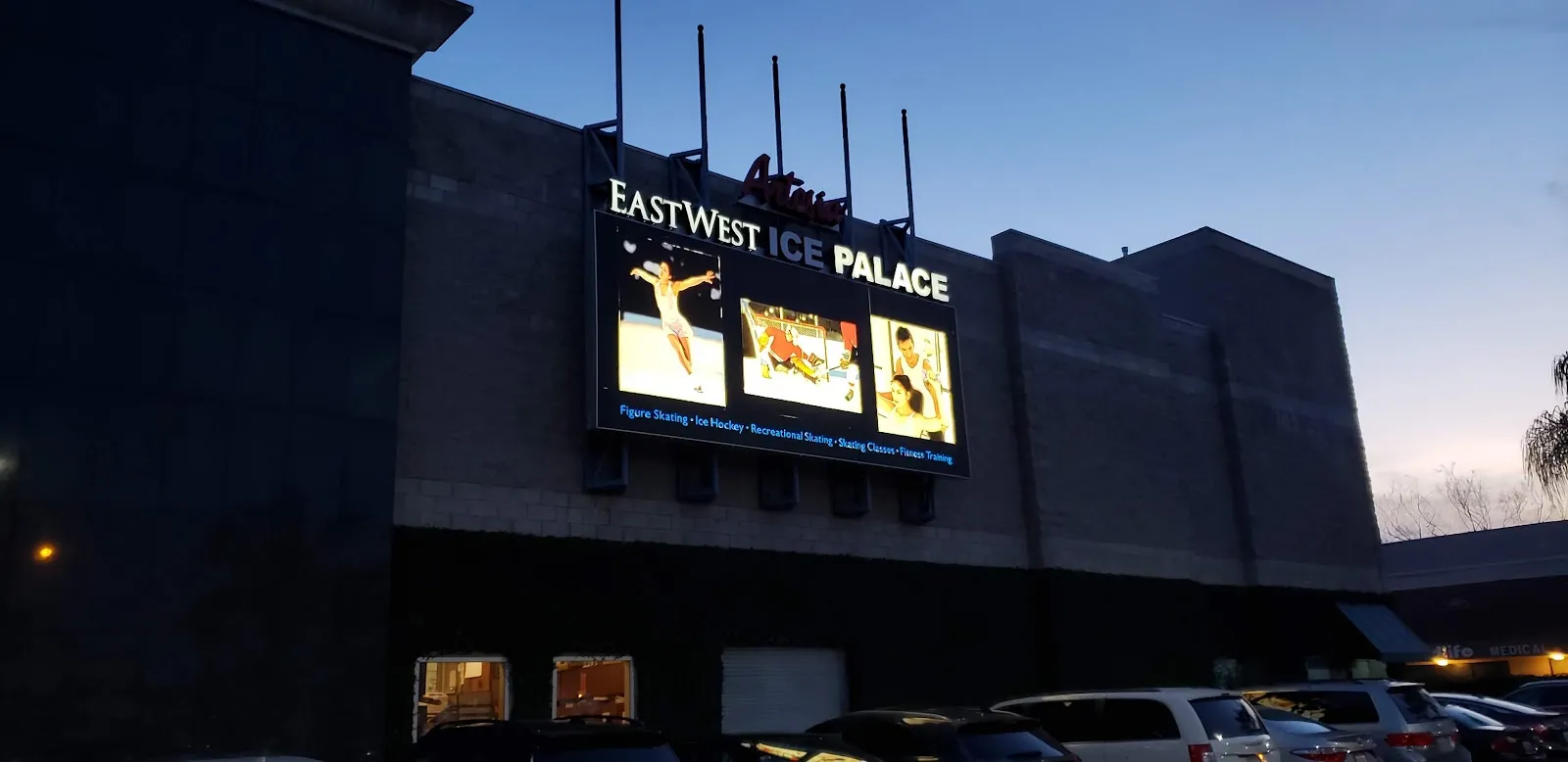 El frente del edificio al anochecer, con su fachada oscura y el letrero iluminado de East West Ice Palace sobre el estacionamiento. — East West Ice Palace