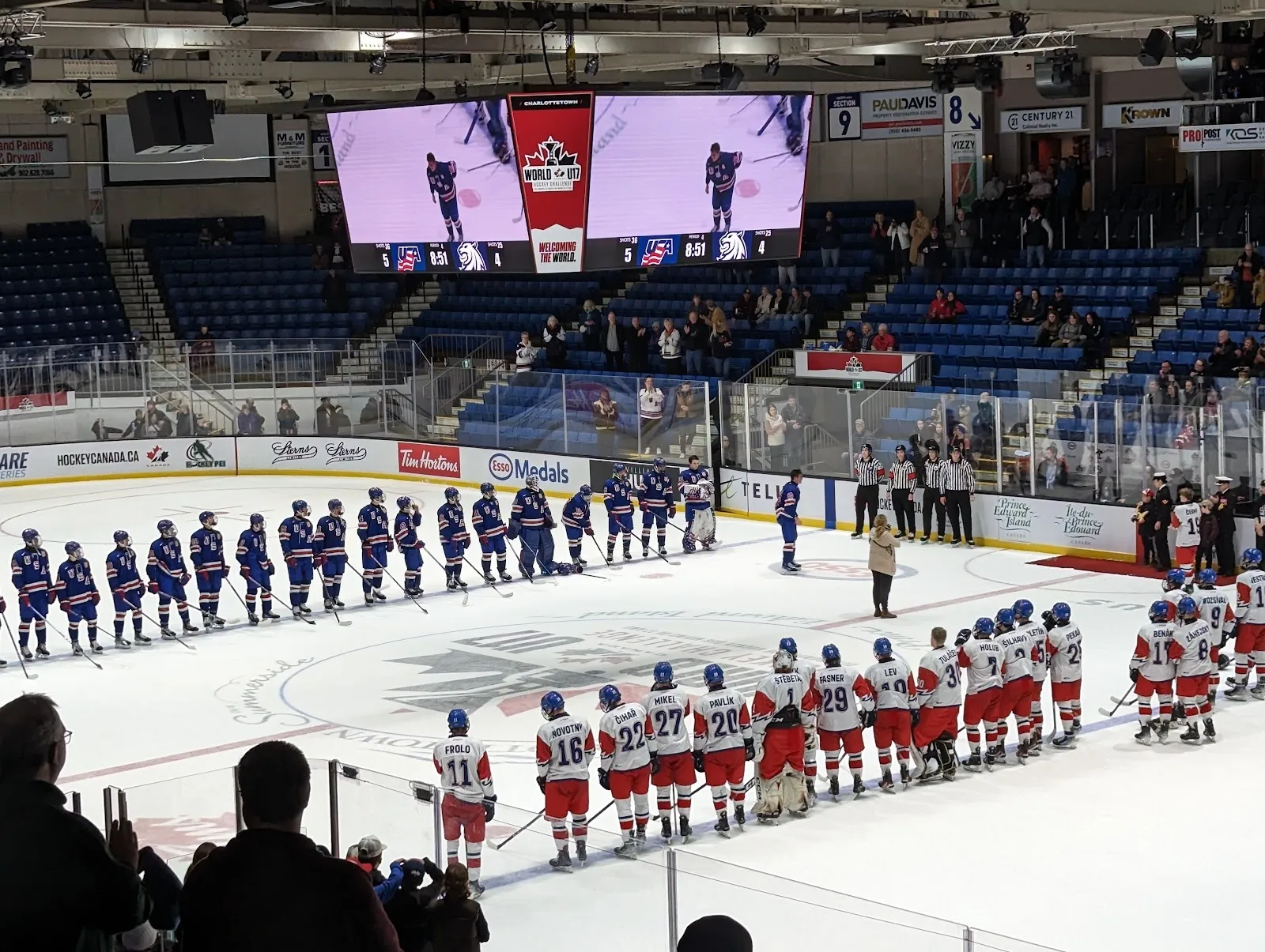 Dos filas de jugadores en las líneas azules de la arena antes de un partido, con asientos rojos y azules alrededor de la pista y marcadores arriba. — Eastlink Centre