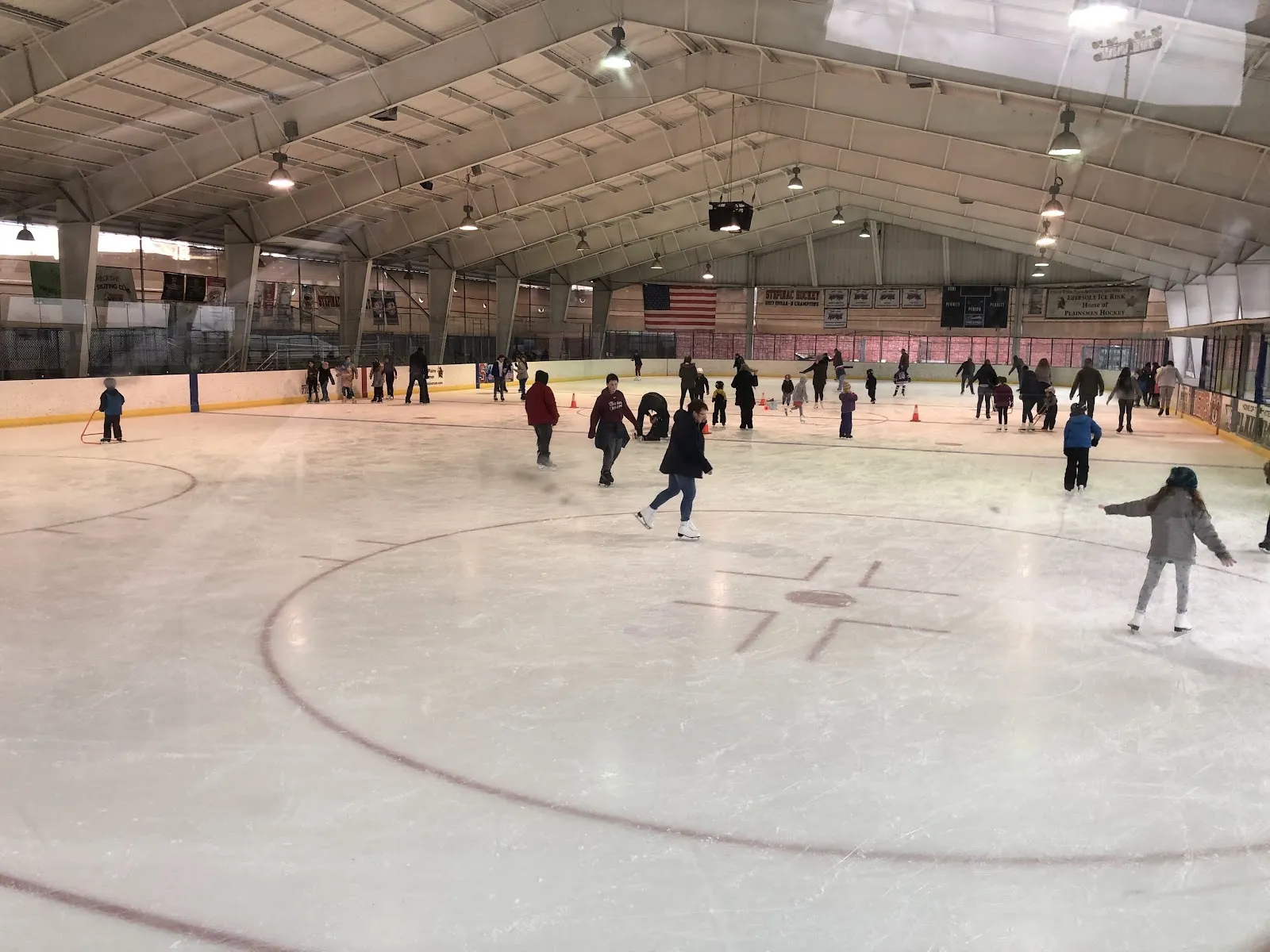 A public session on a wide sheet under a long covered roof, skaters spread across the ice around a centre circle. — Ebersole Ice Rink