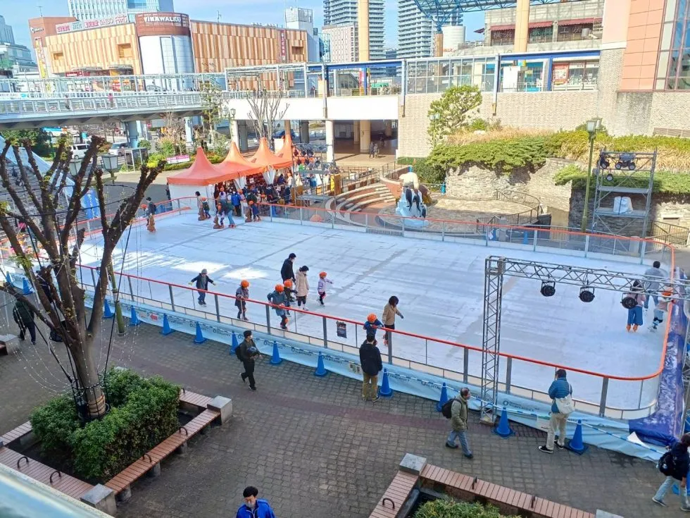 The rink in the Ebina plaza seen from a raised walkway, with distant skaters and the shopping towers behind. — Ebina Central Park Wakuwaku Ice World