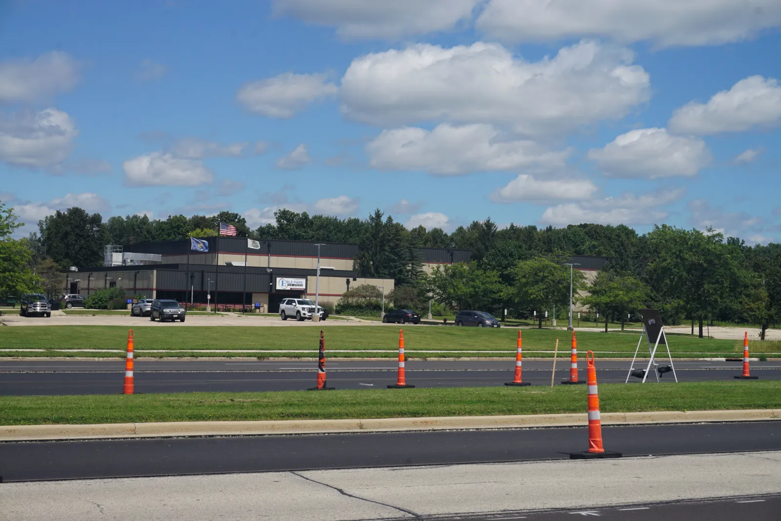The tan-and-black metal-clad arena building seen across the parking lot and access road, with a lit "Eble Park Ice Arena" sign, a US flag and a Wisconsin flag on the roofline, trees behind, and traffic cones in the foreground. — Eble Park Ice Arena