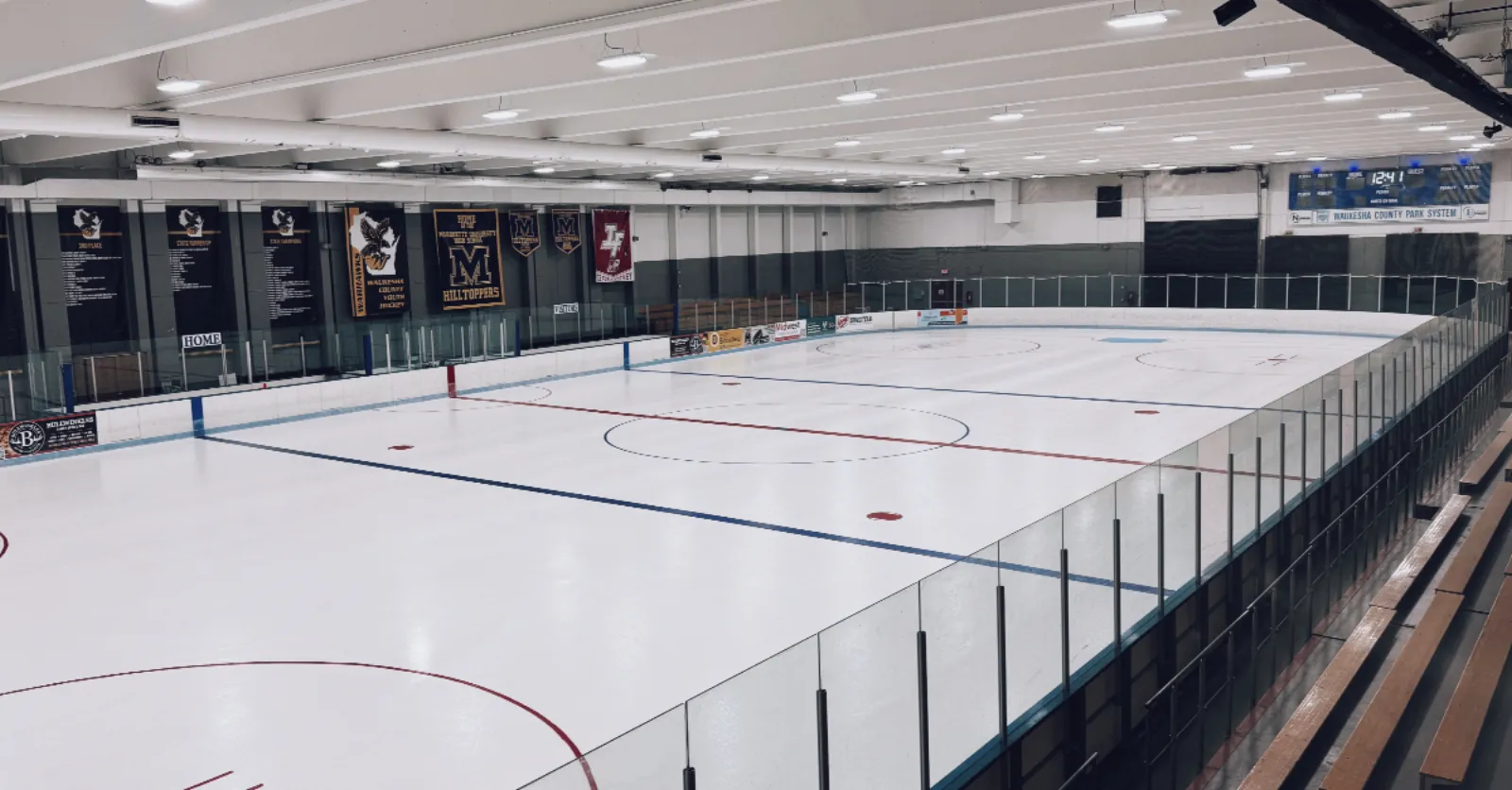 Empty NHL-size ice sheet from the stands, showing rink boards, wooden bleachers, championship banners for local youth and college hockey teams, and a scoreboard reading Waukesha County Park System. — Eble Park Ice Arena