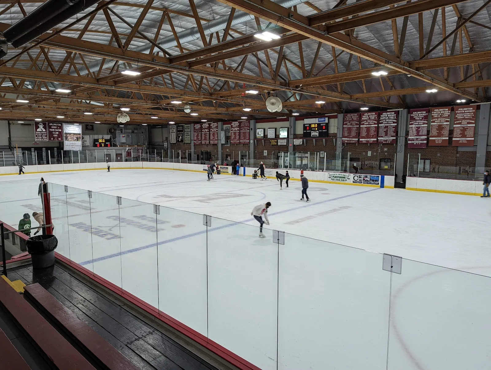 The ice at Ed Burns Arena in Arlington seen from the stands under its timber roof trusses, with a few skaters spread across the sheet and the team banners on the far wall — Ed Burns Arena