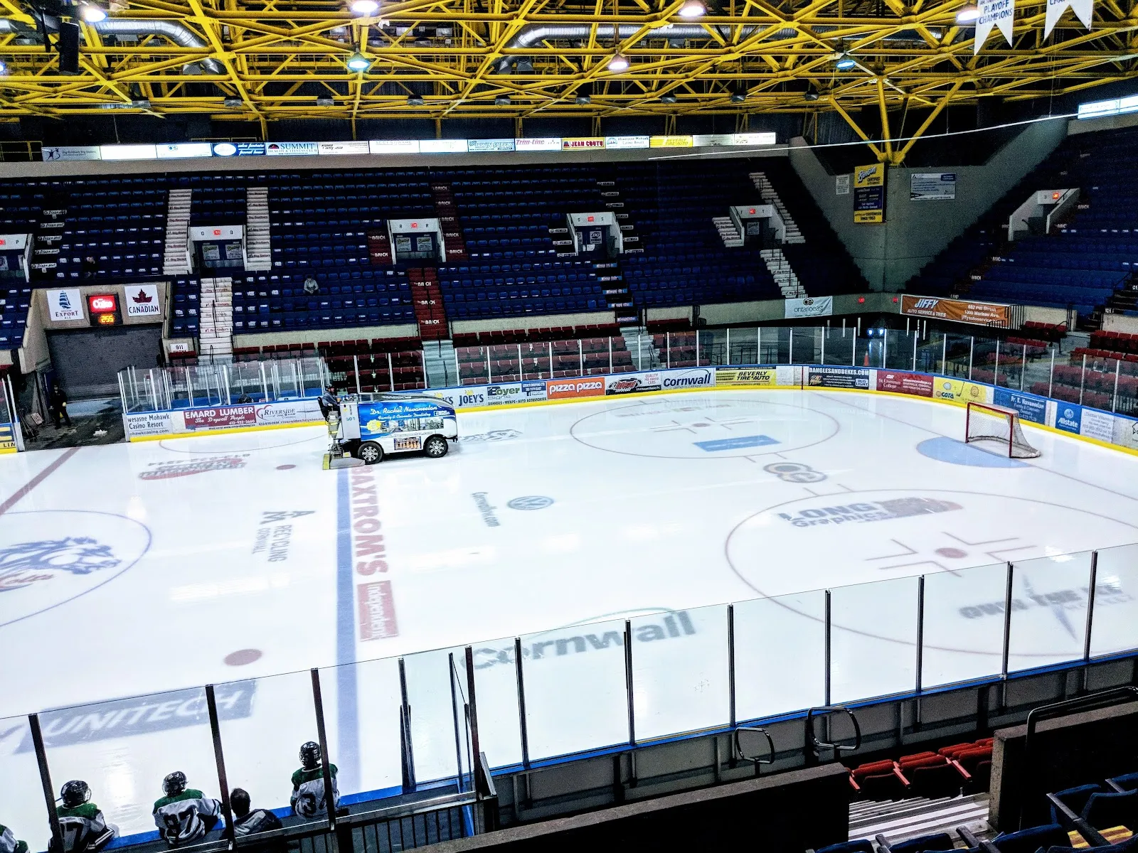 The arena bowl from the upper seats: the ice with an ice resurfacer at centre, blue seating on three sides and a yellow steel roof. — Ed Lumley Arena (Cornwall Civic Complex)