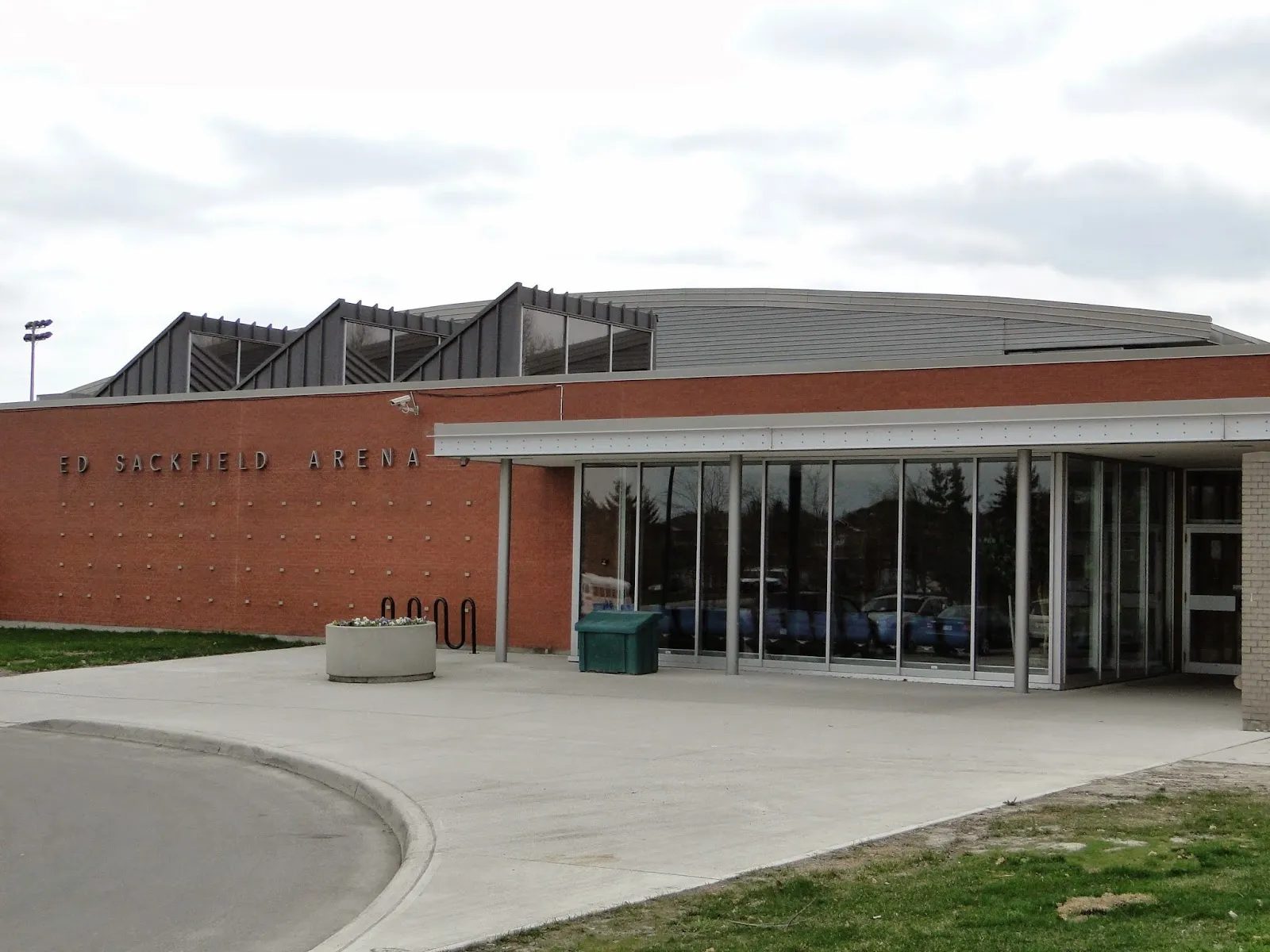 La fachada de ladrillo de la entrada con el nombre de la arena en el muro, un vestíbulo de vidrio y el techo curvo de la arena detrás. — Ed Sackfield Arena (Richmond Hill)