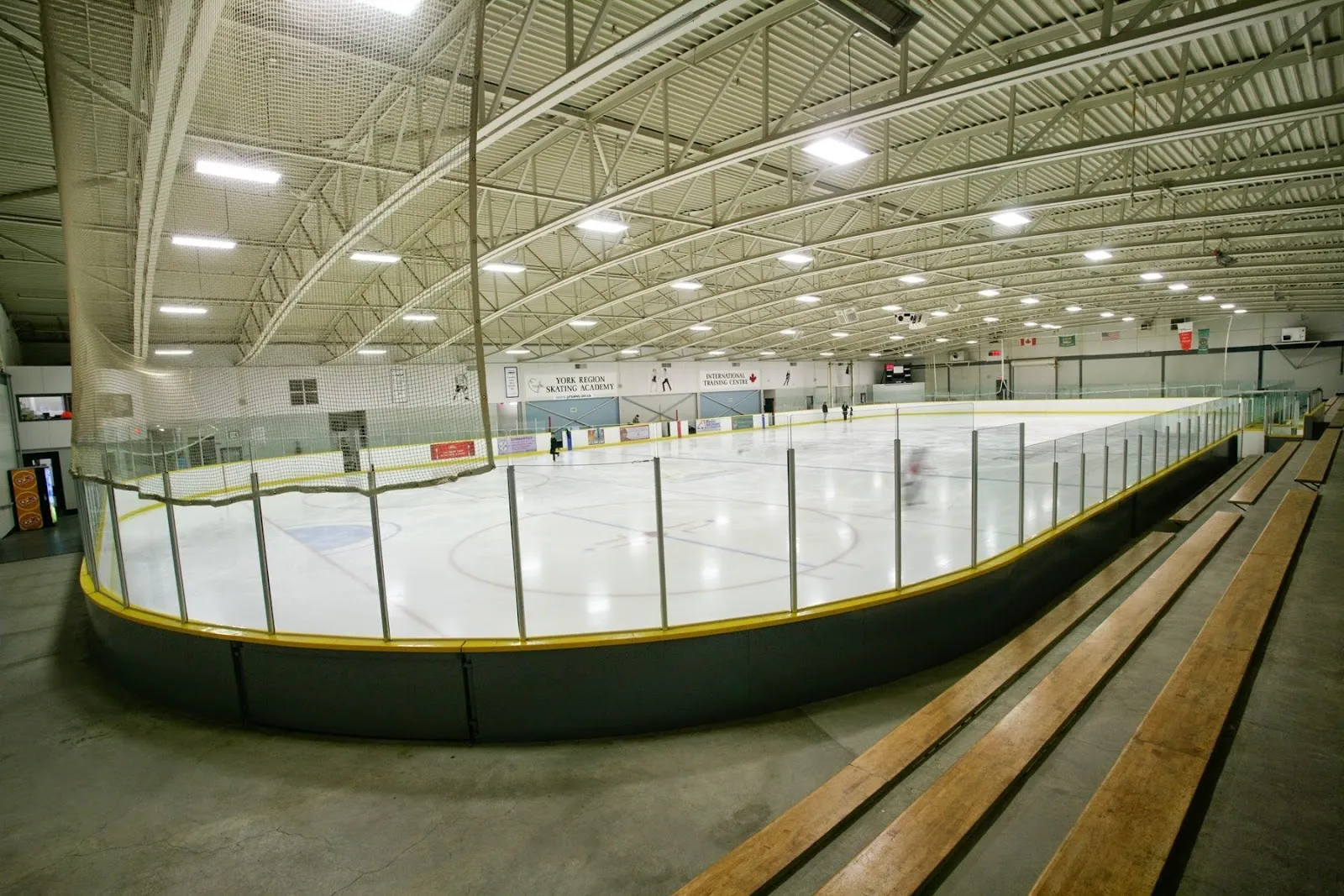 Una vista amplia a lo largo de una banca de madera de la pista, con vallas curvas de ribete amarillo bajo un techo de armaduras de acero verdes. — Ed Sackfield Arena (Richmond Hill)