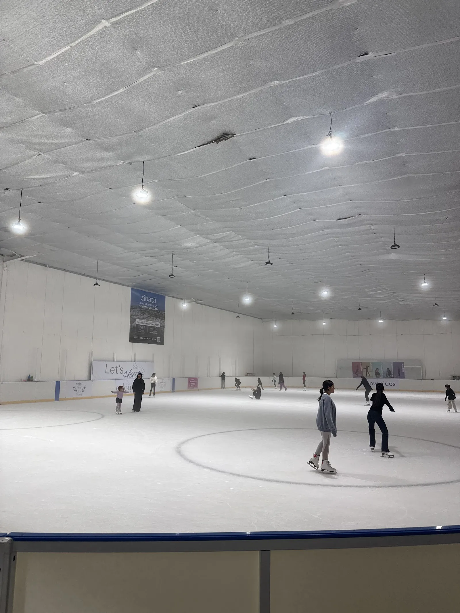 Ice sheet in a white hall under ceiling lights, with a few skaters near centre ice. — Eden Ice