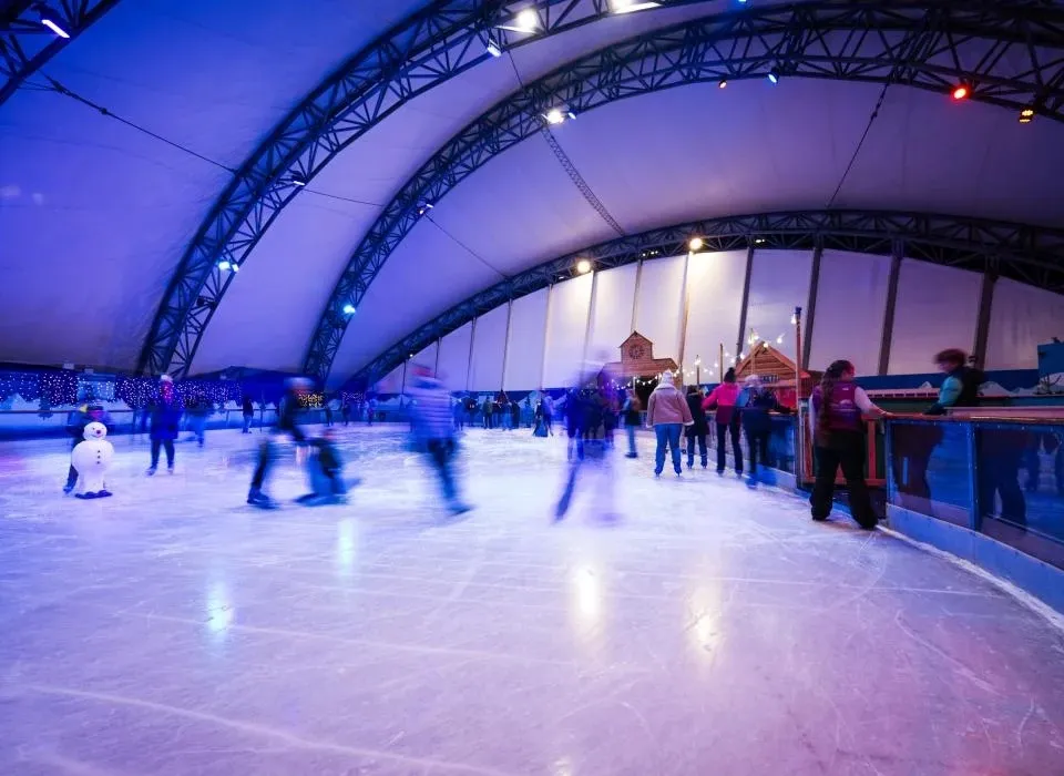 Skaters on the ice under the arched blue and purple tent roof of the Eden Project rink, with the rink-side barrier and wooden chalets behind. — Eden Project Ice Rink