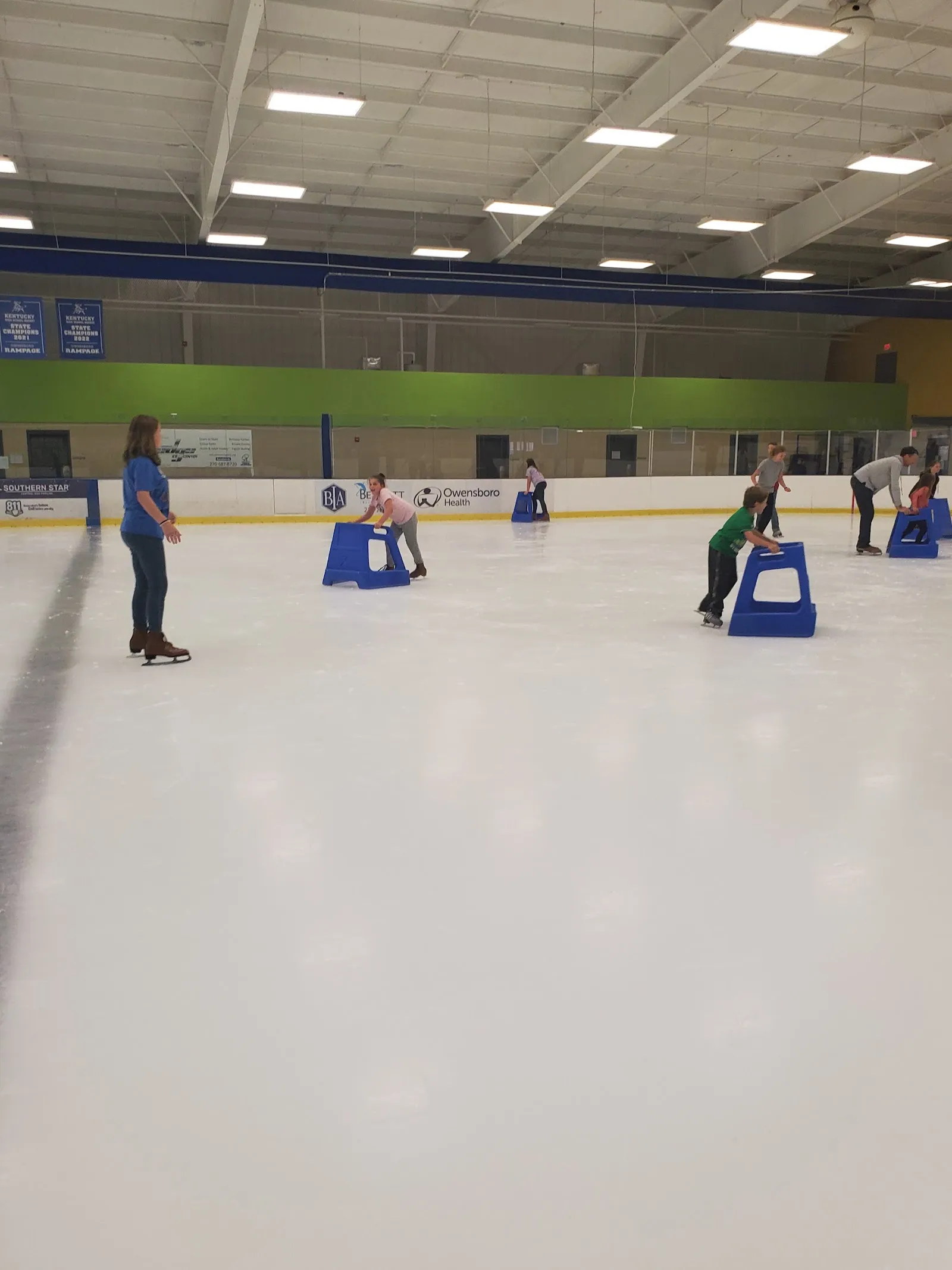 Empty-looking ice seen from the end of the sheet, a few blue skating-aid sleds and a couple of people far down the ice under a lit ceiling with green walls behind. — Edge Ice Center