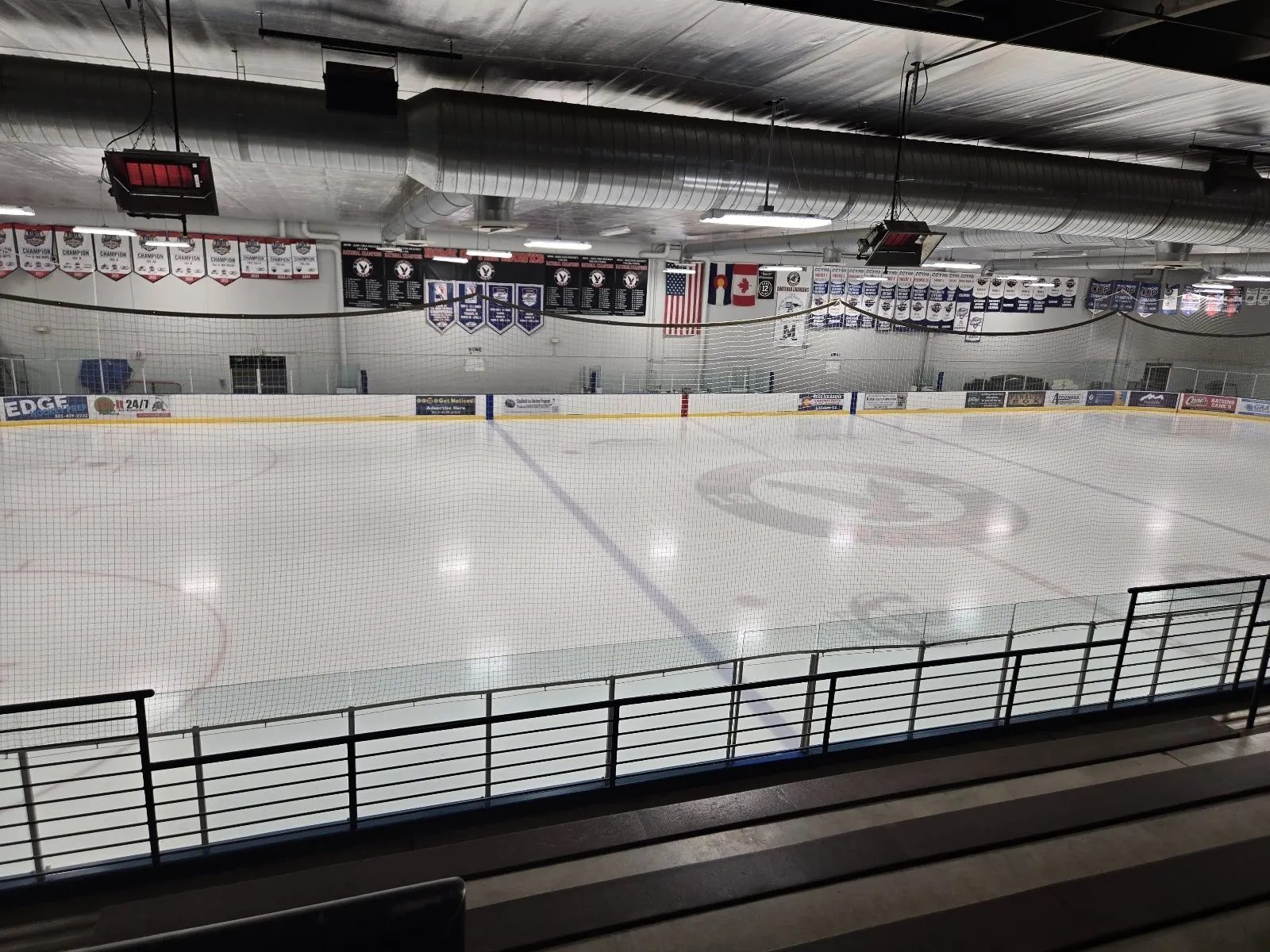 Una vista amplia de la pista por encima de un barandal negro, con hielo blanco y un círculo de saque bajo un techo con banderines de equipos y un marcador. — Edge Ice Arena