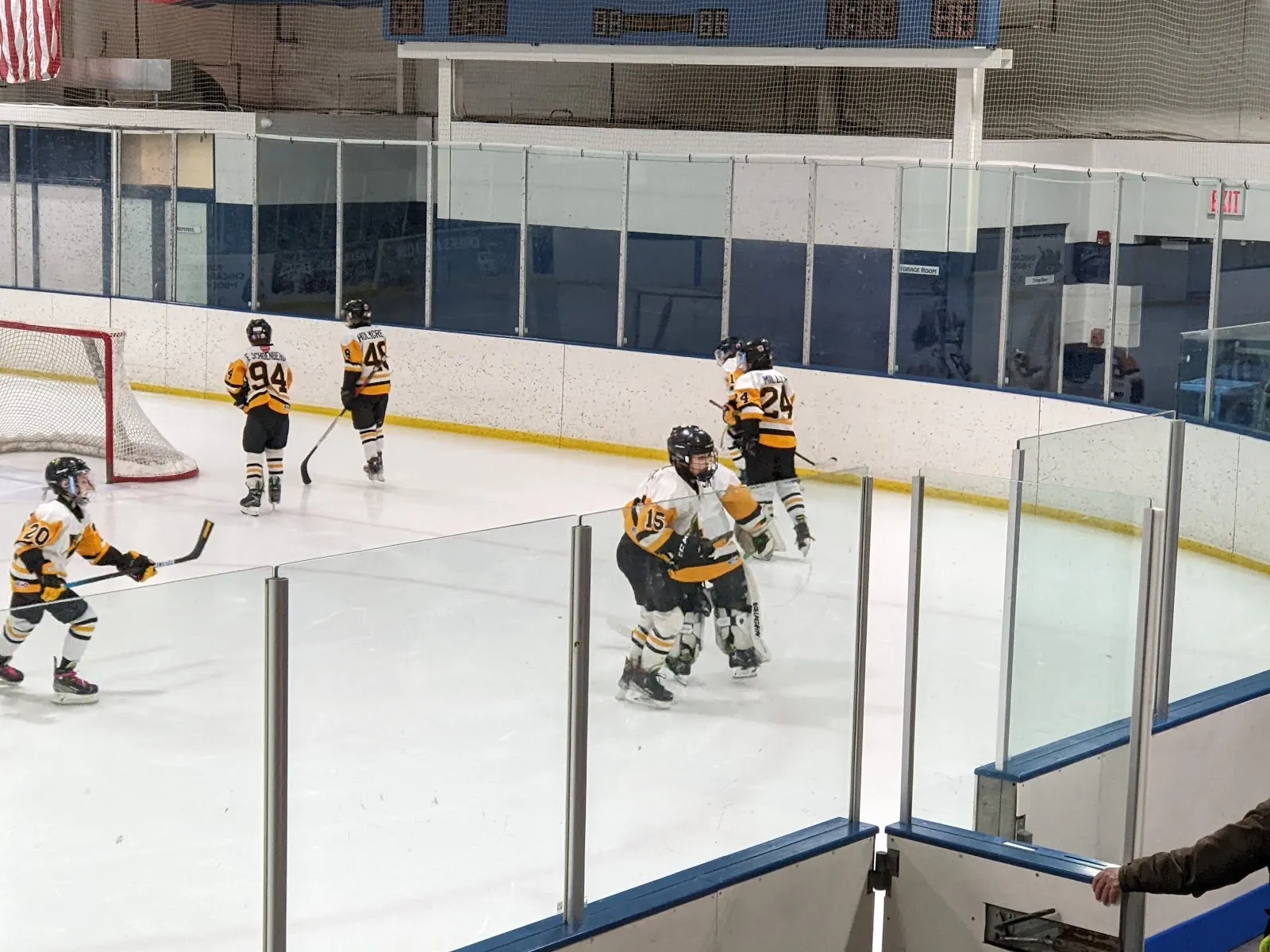 Players spread near the near boards of the sheet seen through clear glass panels, with a goal at the left and sponsor boards along the wall. — The Edge on John St (Bensenville)