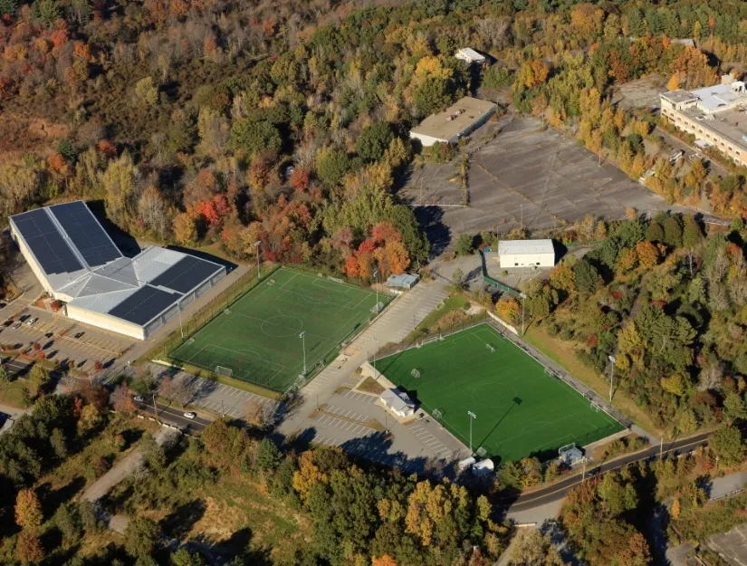 Vista aérea del Edge Sports Center: el edificio de la pista con su techo de paneles solares junto a los dos campos de pasto sintético, rodeado de árboles otoñales. — The Edge Sports Center