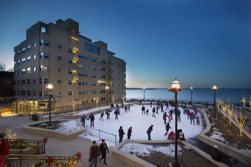 The Edgewater hotel's tall pale stone front at blue hour with the rink in front of it, skaters on the ice and lamps along the lakeside rail. — The Edgewater Ice Rink