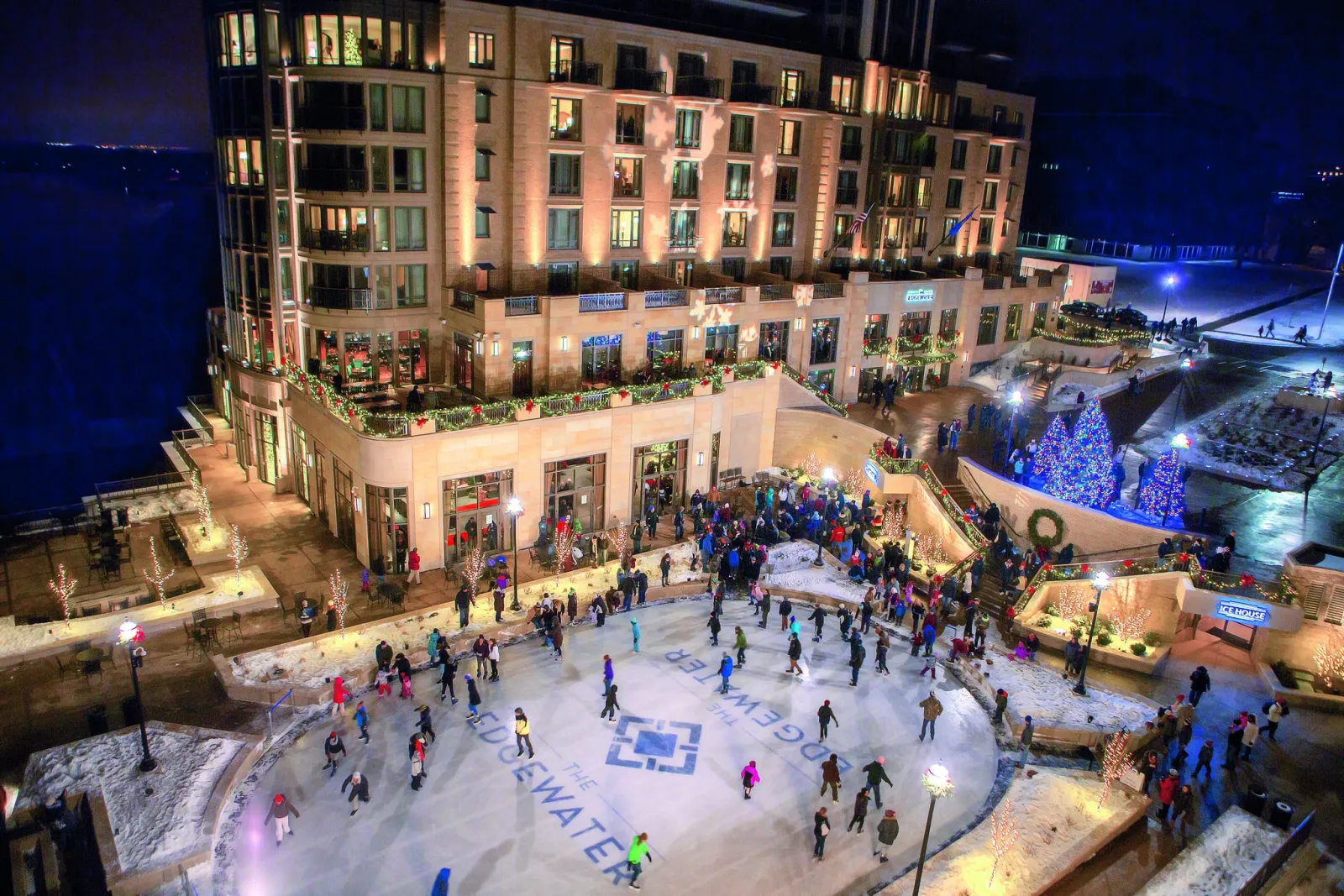 The Edgewater rink at dusk, skaters spread over the ice below the hotel's lit terraces with a Christmas tree and the lake beyond. — The Edgewater Ice Rink