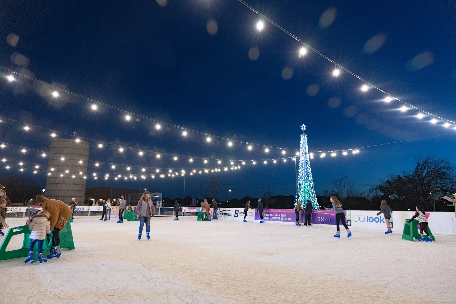 The ice at dusk under strings of lights, with a lit tree-shaped tower and a grain silo beyond the boards and a few skaters, one family pushing a child on a green skating aid. — Edmond Ice Rink