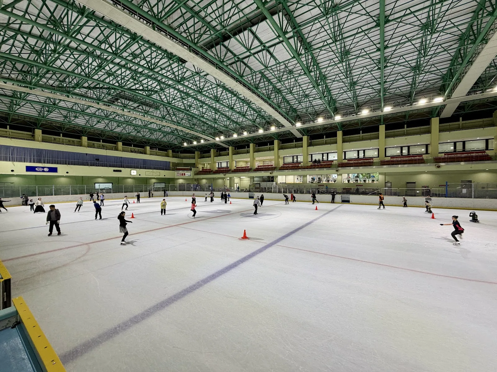 The 60 by 30 metre indoor ice rink at Edogawa Sports Land under a green steel roof, with public skaters spread out across the ice and orange cones in the foreground — Edogawa Sports Land Ice Rink