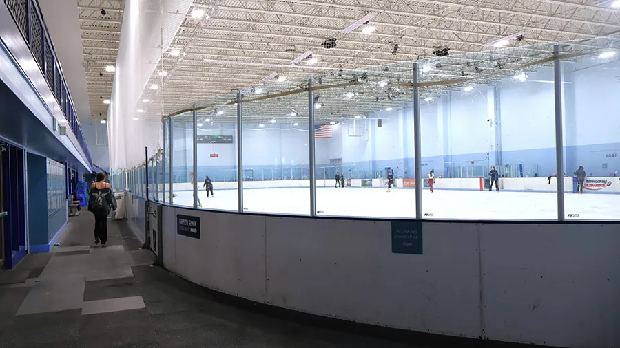 Wide view of the Green Rink ice sheet through the glass from the walkway, with rink boards, an overhead scoreboard, and a few skaters on the ice. — Edora Pool Ice Center