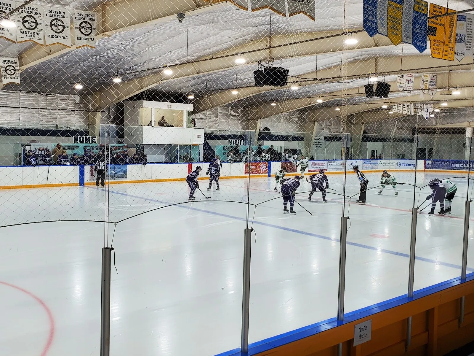 A game under a yellow-banner ceiling seen over a clear rail, players spread across a wide sheet with a blue-lit boards band. — Edson & District Leisure Centre