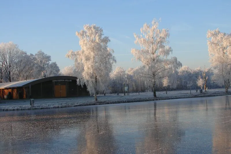 Glad ijs dat berijpte bomen weerspiegelt naast het clubgebouw van Eemnes onder een heldere blauwe winterlucht. – Eemnesser IJsclub De Vaart