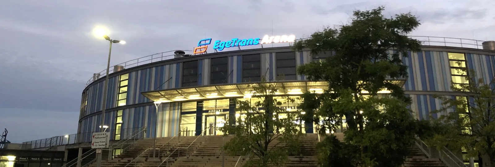Die geschwungene Glas-Stahl-Front der EgeTrans Arena in der Dämmerung, das Dach beleuchtet und das Eingangsschild über Bäumen im Vordergrund. – EgeTrans Arena