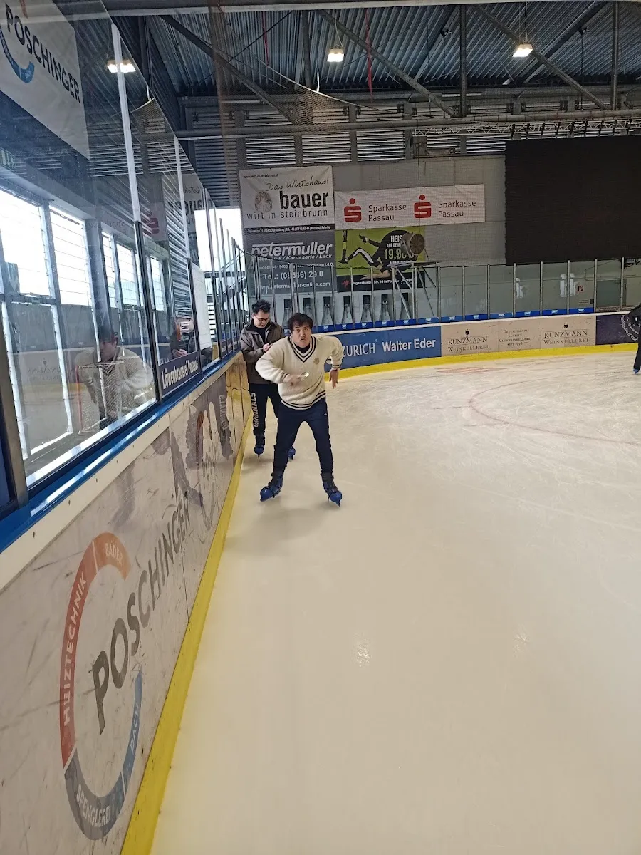Die Eisfläche der Arena von der Bande aus gesehen, mit blau-gelben Eisrändern, Sponsorentafeln an der Wand und einem erwachsenen Schlittschuhläufer am rechten Rand. – Eis-Arena Passau