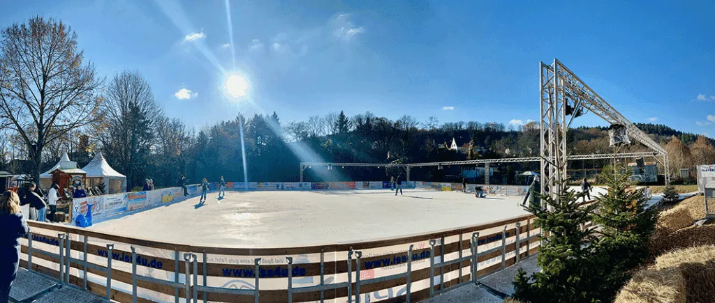 The fenced real-ice rink in winter sun with a few skaters far away, trees and a light gantry beyond the boards. — Eis Eis Wallhausen (Echt-Eisbahn am Freibad)