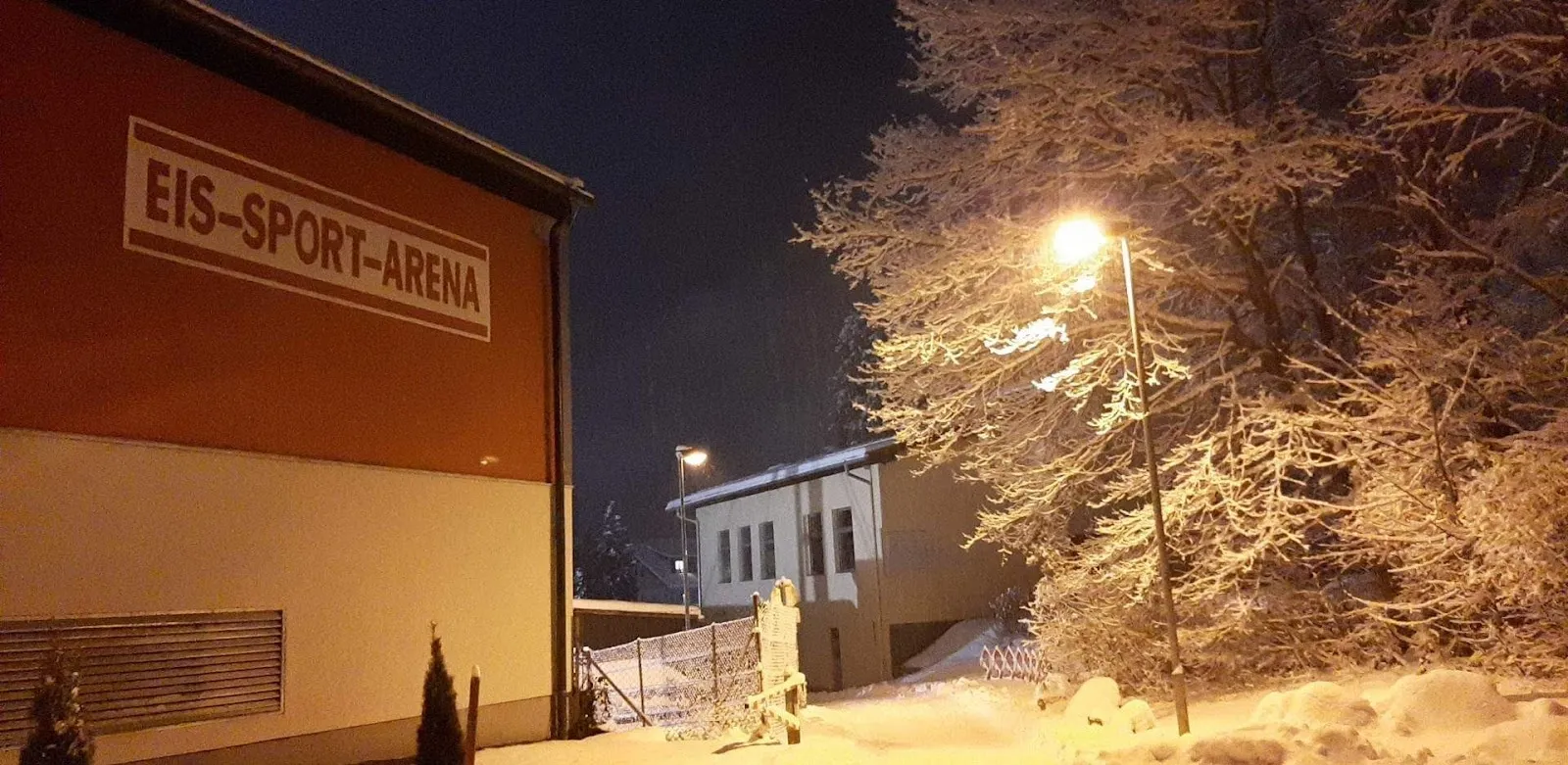 Der Schriftzug „Eis-Sport-Arena“ an der Seitenwand der Eishalle Spittal bei Nacht, daneben verschneite Bäume und eine Straßenlaterne. – Eis-Sport-Arena Spittal