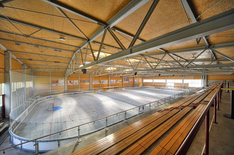 Die leere Eisfläche der Eis-Sport-Arena Spittal, von der Holztribüne aus gesehen, unter einem Dach aus Holzbindern – Eis-Sport-Arena Spittal