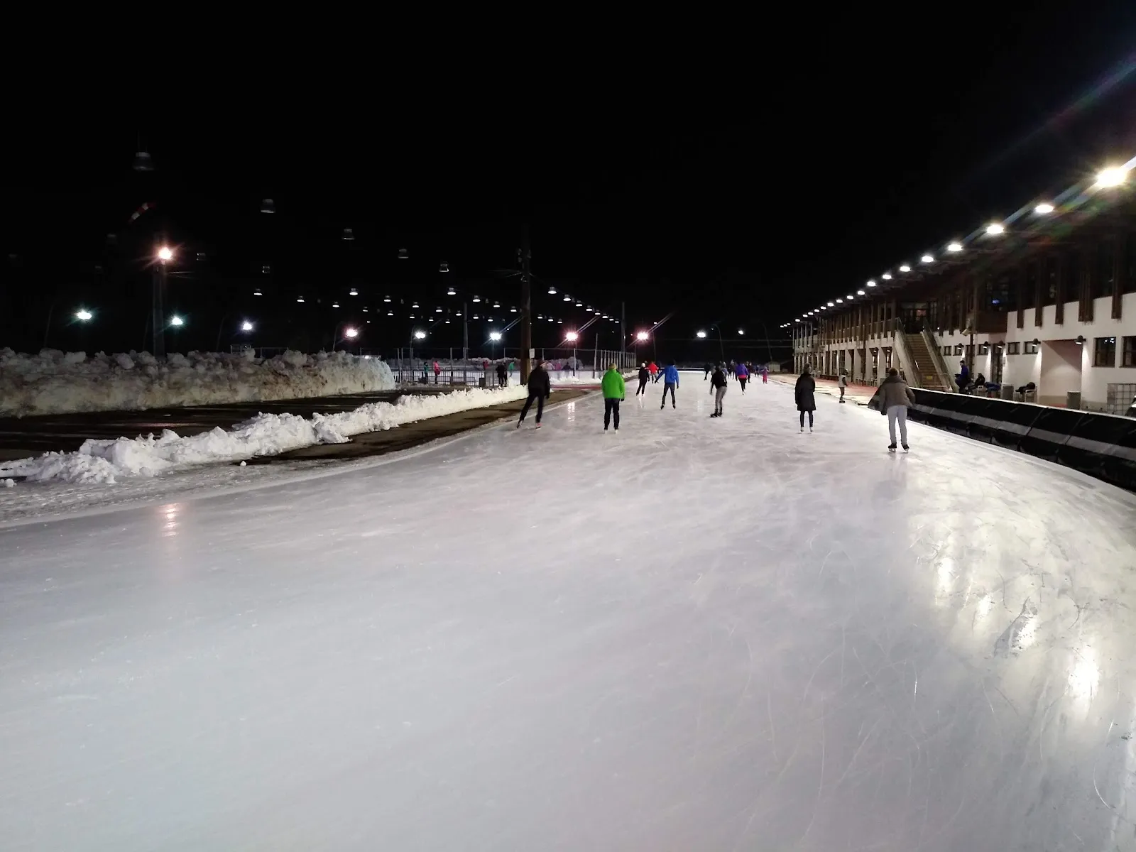 The long curved outdoor ice track under floodlights at night, snow heaped along the inside and a few skaters far down the straight. — Städtisches Eis- und Funsportzentrum Ost